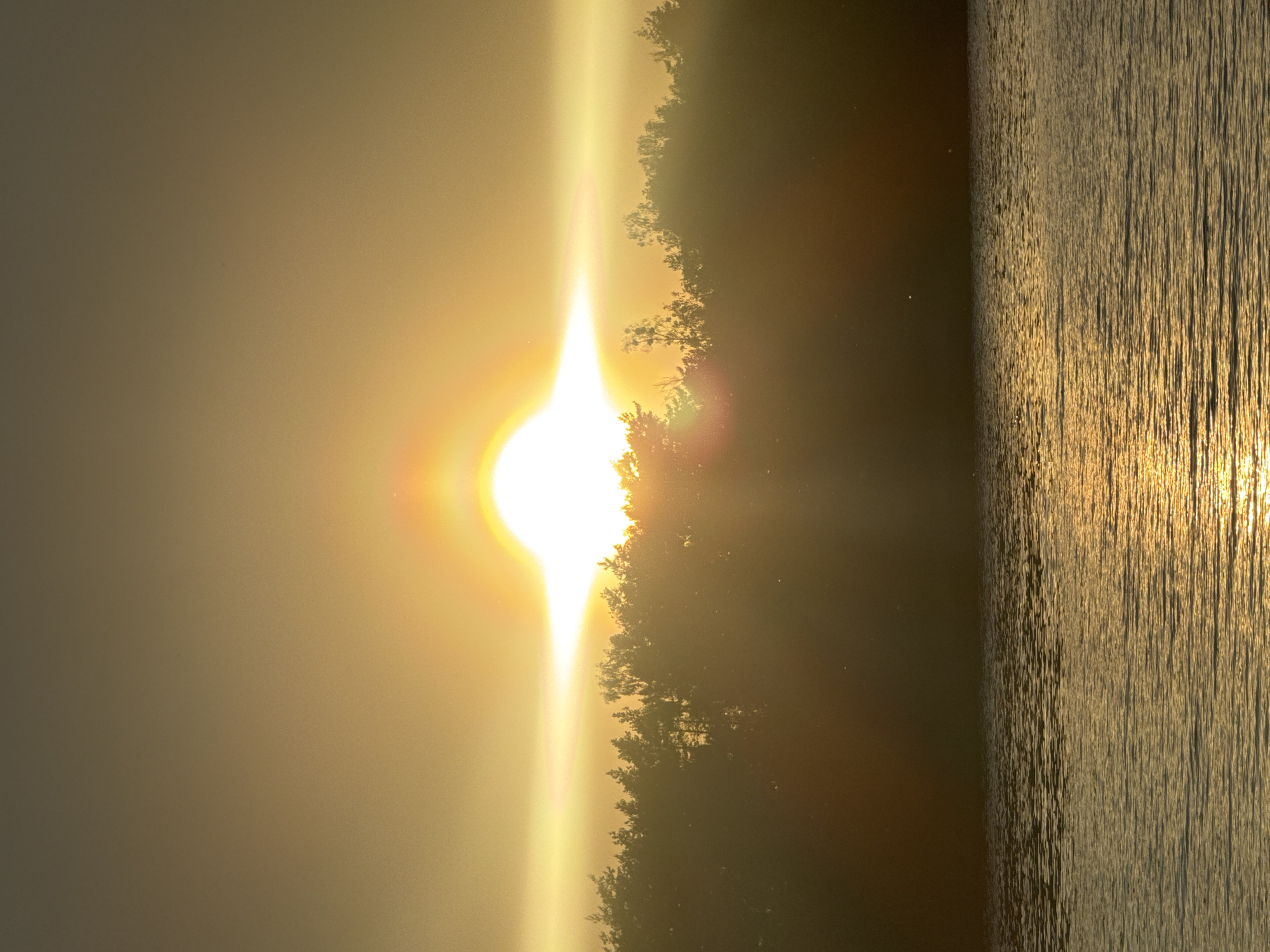The image depicts a horizontal view of what appears to be a sunset over a body of water, possibly a lake or river, in the vicinity of Абухаўскі сельскі Савет, Belarus. The bright sun is partially obscured by a line of trees or vegetation along the shoreline, creating a strong glare. The surface of the water reflects the golden sunlight, with vertical striations suggesting ripples or small waves. The left side of the image shows a hazy or cloudy sky.