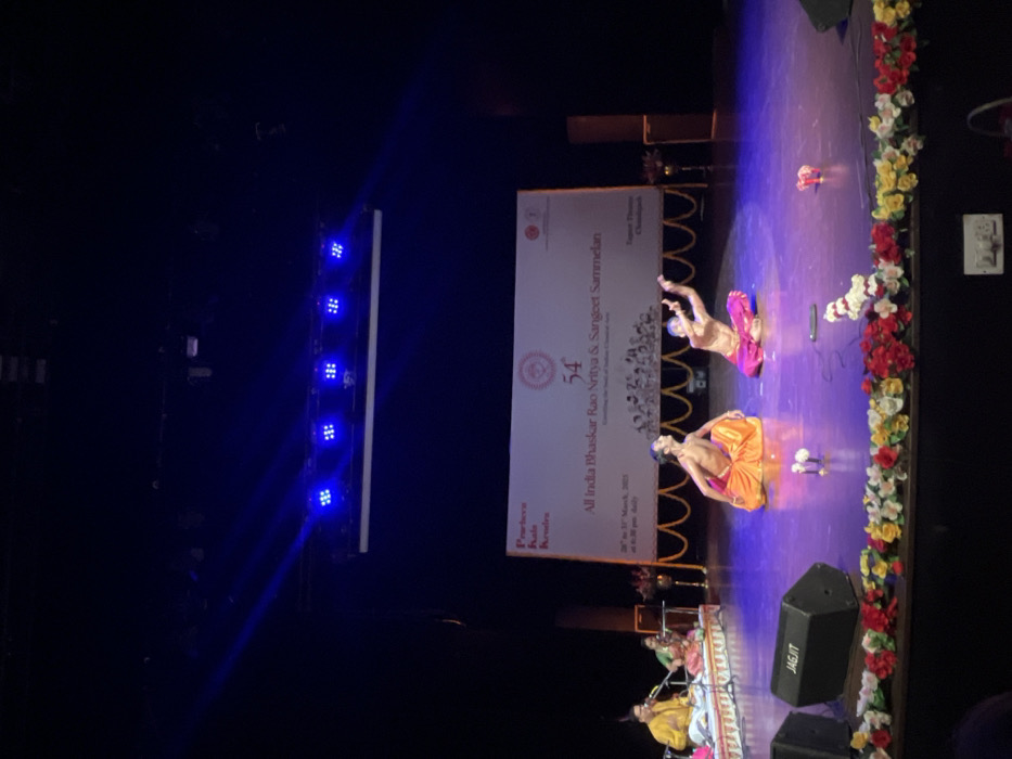 The image shows two men in orange and red traditional Indian clothing performing a dance on a stage. The stage is decorated with flowers and has a white banner that says "54th All India Bhaskar Rao Nritya & Sangeet Sammelan". The event took place at the Tagore Theatre in Chandigarh, India, located at latitude: 30.733, longitude: 76.789. There are two additional men in the background playing instruments. Above the stage is a row of blue LED lights. The stage is illuminated with blue spotlights. The background appears to be dark and empty except for the banner and a speaker on the left side of the stage. The image is taken from the audience area, giving a slightly elevated view of the stage.