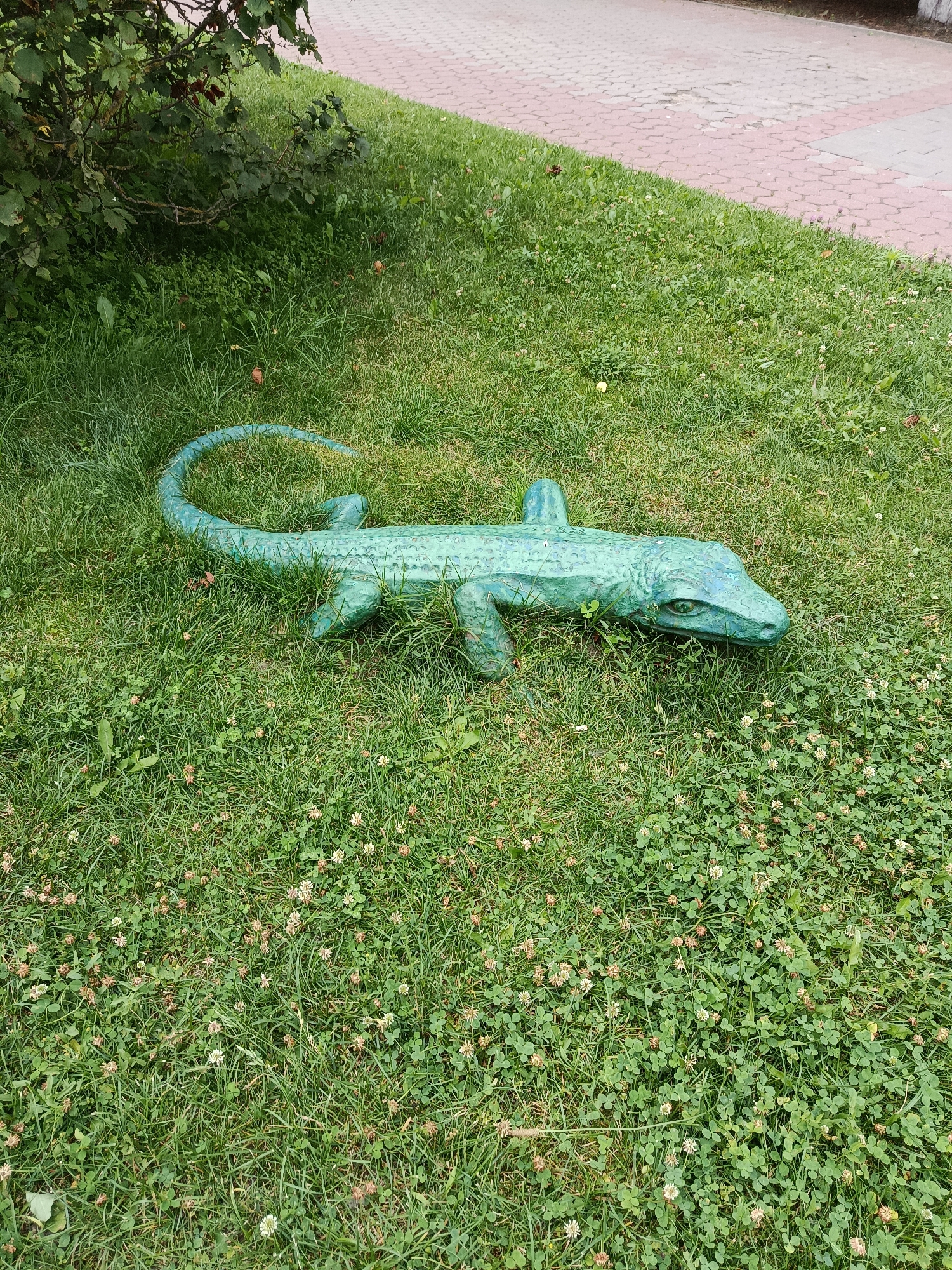 In Zhlobin, Belarus, a green reptilian sculpture lies on a grassy area next to a paved walkway. The sculpture, possibly representing a lizard or alligator, is positioned horizontally in the grass, its body extending from the upper left to the lower right of the frame. Its tail curls upwards, and its head, facing towards the right, has visible eyes and snout. The grass is a vibrant green, dotted with clover flowers. The paved walkway, composed of hexagonal-patterned bricks in shades of red and grey, runs diagonally across the upper portion of the image. A bush with green leaves is visible in the upper left corner. No people are present in the scene.