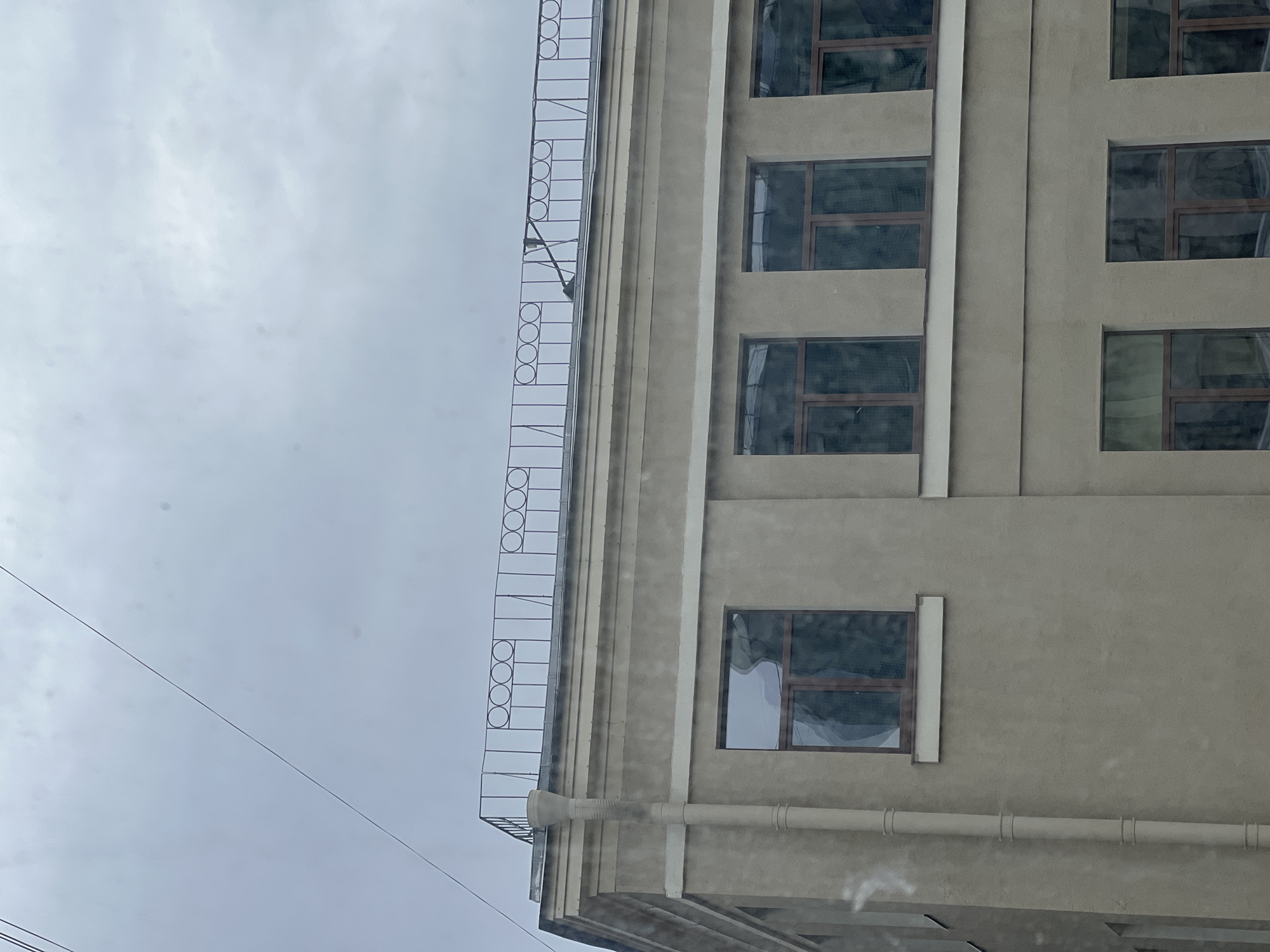 The image depicts the corner of a building in Saint Petersburg, Russia. The building is constructed with a beige facade and features rectangular windows with dark frames. A metal fire escape runs vertically along the corner of the building, extending from the top towards the bottom. The sky above is overcast with grey clouds. A power line stretches diagonally across the left side of the image. A drainpipe follows the exterior of the building.