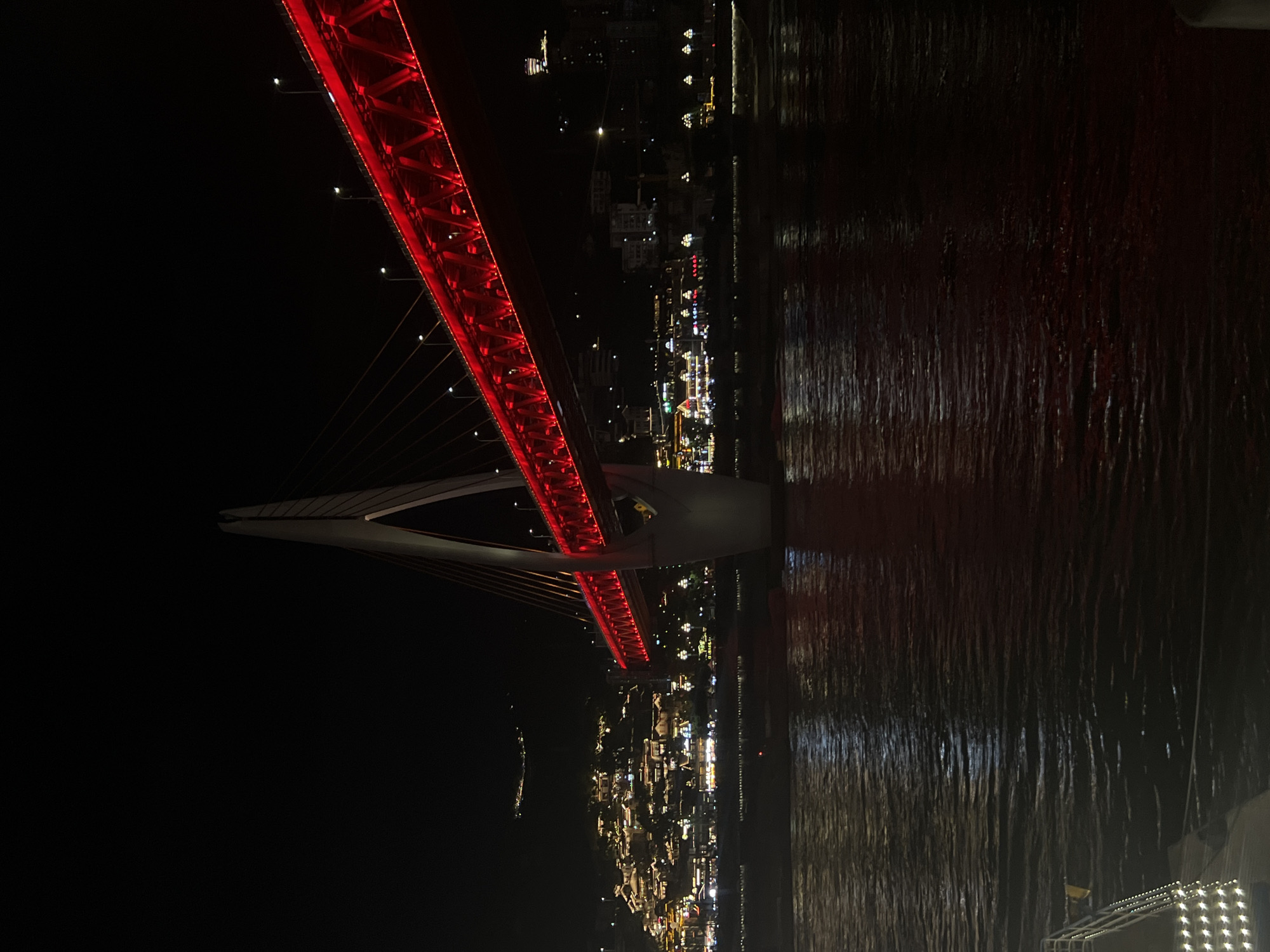 A night view of the Hongyadong Bridge in Chongqing, Yuzhong District, China, showcases the illuminated structure spanning a body of water. The bridge is prominently lit with vibrant red lights along its deck, creating a striking visual against the dark sky. The bridge's main support tower, a white A-frame structure, is also partially visible. In the background, the city skyline of Hongyadong is illuminated with numerous warm-toned lights, indicating buildings and structures on a hillside. The water in the foreground reflects the lights from the bridge and the city, creating a shimmering effect. The scene is devoid of any people or vehicles, focusing solely on the architecture and the nighttime ambiance.