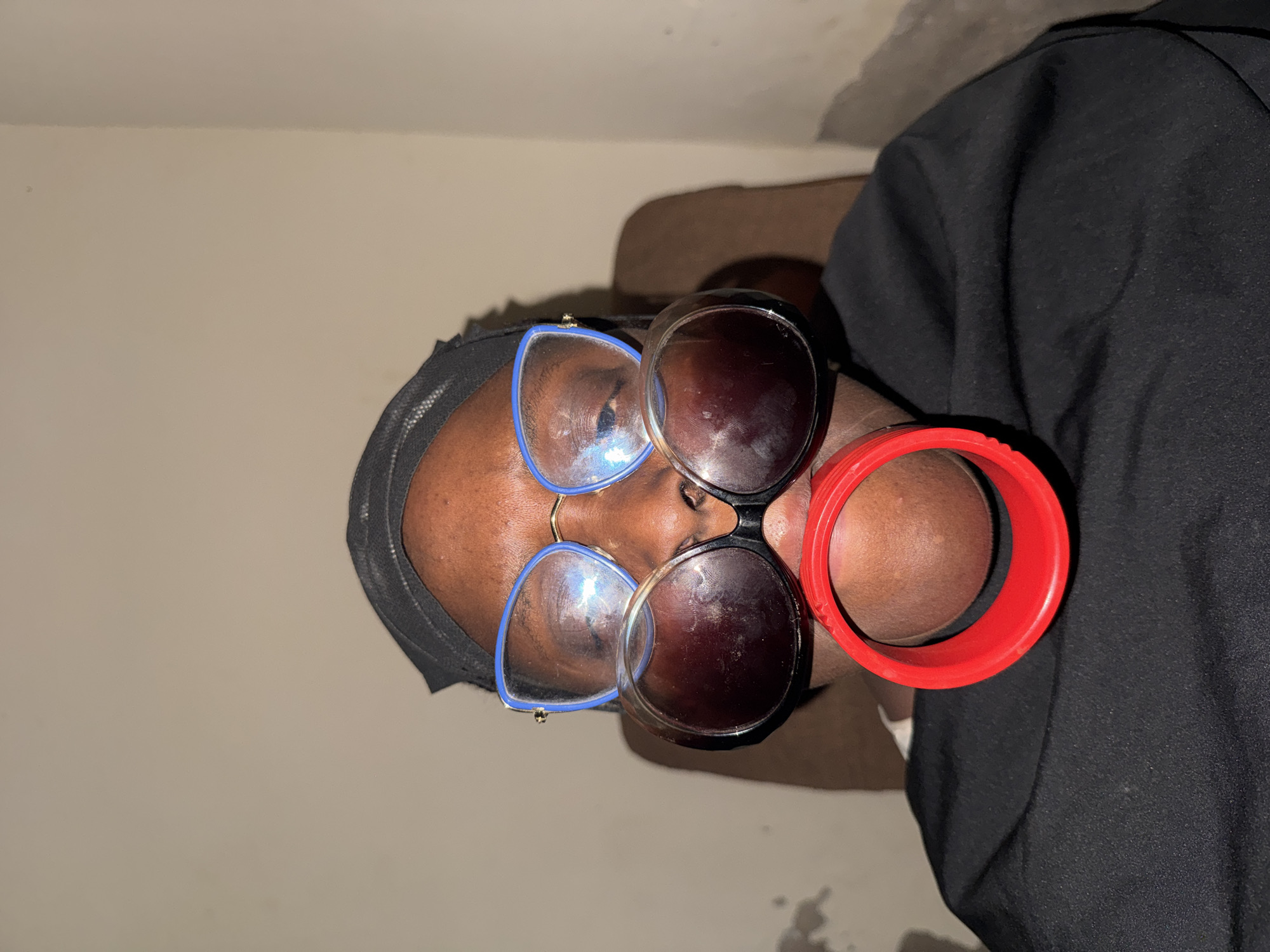 A close-up, rotated portrait image shows a person with dark skin. The individual is wearing a black head covering and a black garment, possibly a shirt or tunic. Two pairs of eyeglasses are worn simultaneously on their face: one pair features blue frames with clear lenses, and the other has dark frames with dark, tinted lenses, positioned slightly below the first pair. A vibrant red, wide, circular cuff or bracelet is held up to the left side of the person's face, partially obscuring it and their mouth. The person appears to be posing for the camera, with a direct gaze.
The background consists of a plain, light beige or cream-colored wall, with a small portion of a brown padded surface, likely a piece of furniture like a sofa or headboard, visible behind the person's head and shoulder. The lighting appears to be artificial, casting some shadows. No specific time of day or weather can be determined from the image, but the setting is indoors. There is no visible text in the image. The location context provided is Yelwa, Nigeria.