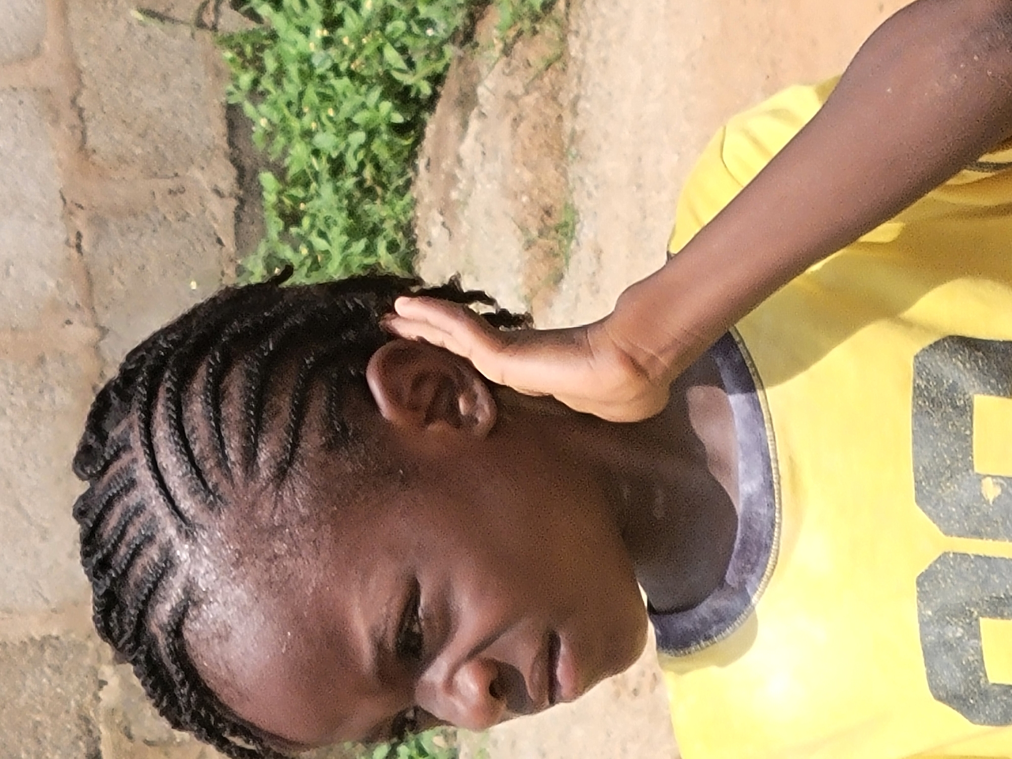 A close-up shot depicts a person with dark skin and braided hair. The individual is wearing a yellow shirt with dark blue trim and the number "6" visible on the front. The person is outside in an area with a concrete or stone surface and greenery growing nearby. The exact location in Madalla, Nigeria, cannot be determined from the image. The individual has their right hand next to their ear.