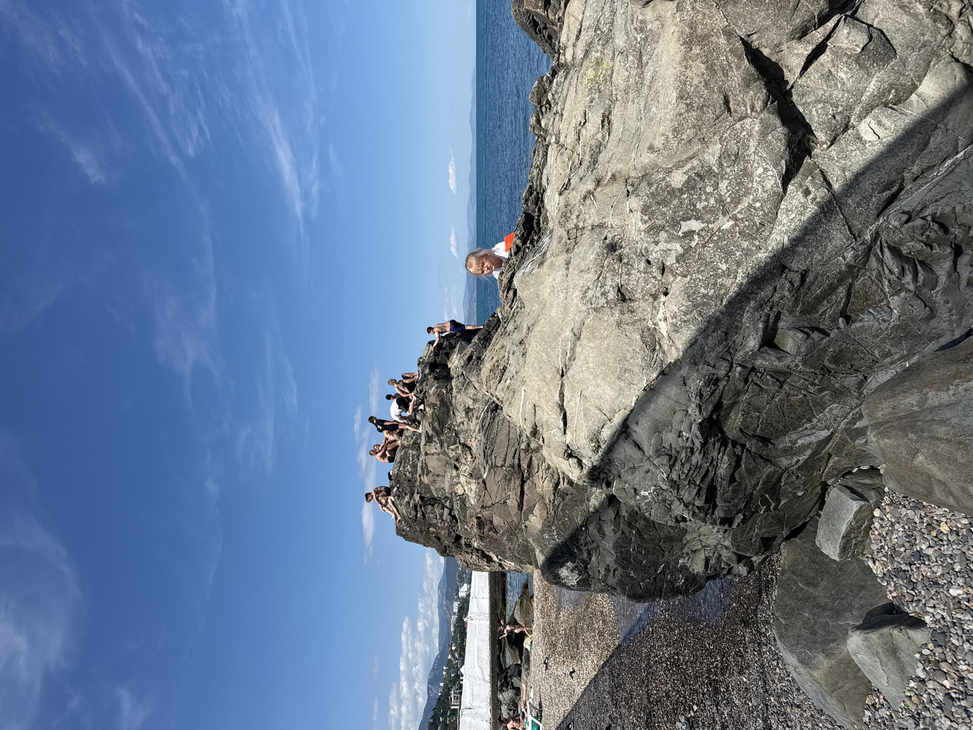 A group of people are sitting on the edge of a large rock formation overlooking the sea. The sun is shining brightly, casting a shadow on the rock. The sky is blue with a few scattered clouds. There are some buildings visible in the distance. The people appear to be enjoying the day, with some of them looking out at the water. The scene appears to be a popular spot for swimming and sunbathing.