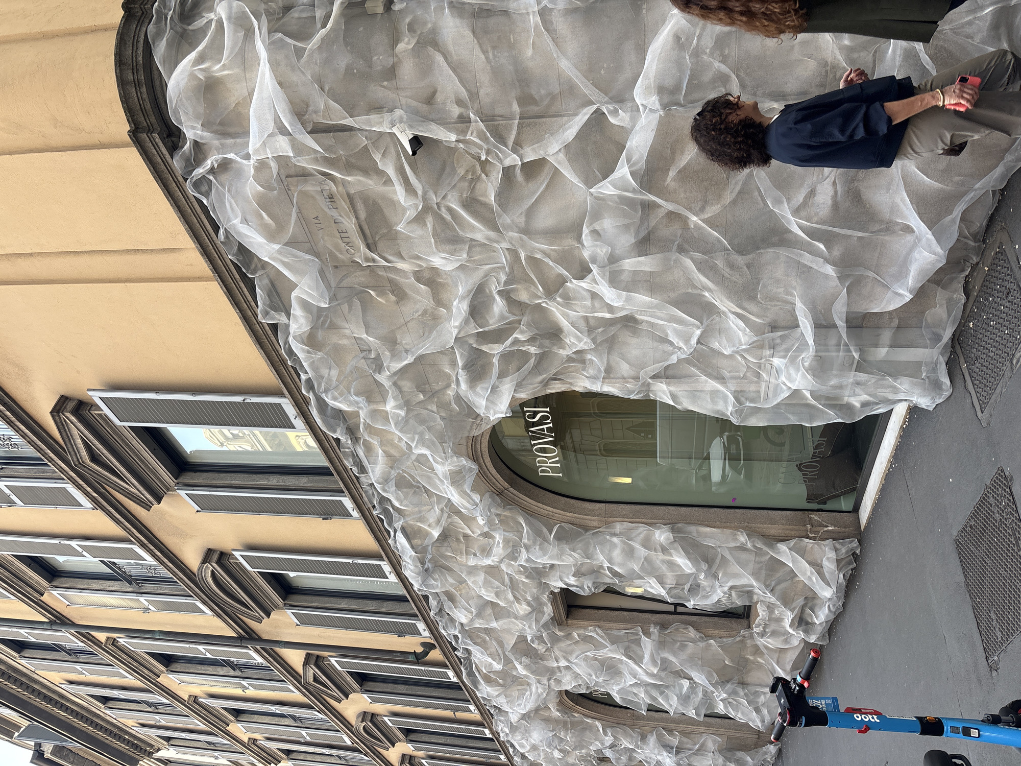The image displays an urban street scene in Milan, Italy, featuring a building facade partially covered by a translucent, crinkled white mesh fabric. This sheer material drapes over the lower and middle sections of the building, creating a soft, sculptural, and cloud-like effect over the stone facade, extending down to the sidewalk.
At ground level, an arched shop entrance is visible, with the word "PROVASI" reflected in reverse on its glass. Above this, a stone plaque on the building clearly displays the street name: "VIA MONTE DI PIETA'". The uncovered upper floors of the building reveal a light yellowish-beige facade with multiple rectangular windows fitted with dark louvered shutters.
Two individuals are present on the sidewalk to the right. One person, with short, curly brown hair, is seen from the side, wearing a dark blue top and light khaki trousers, holding a bright pink smartphone. They appear to be observing the building or passing by. Another person is partially visible behind them. In the lower right foreground, a blue and red electric scooter, branded "oott," is parked on the grey sidewalk next to a metal grate. The scene is set during the daytime, under what appears to be diffused or indirect light.