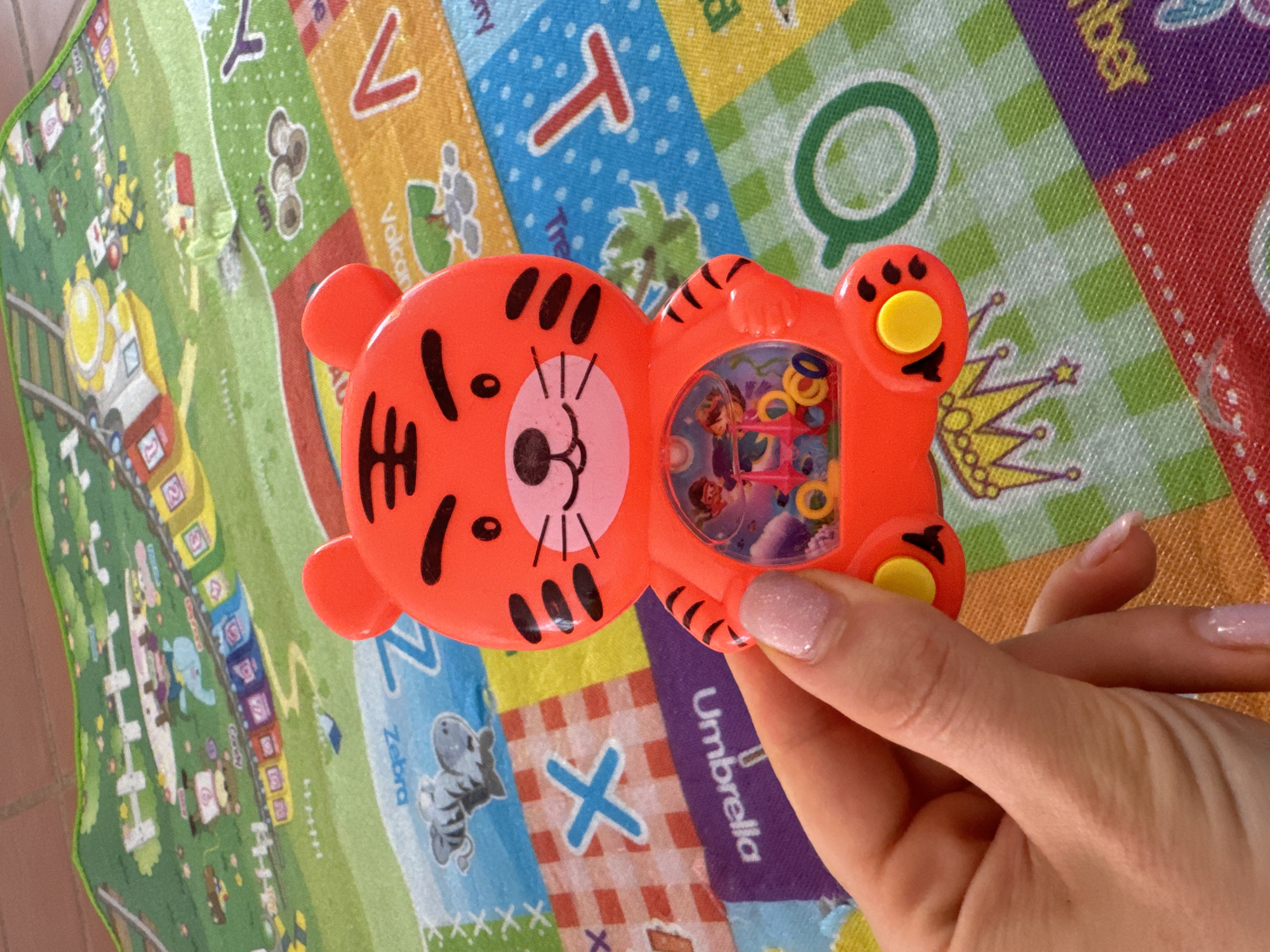 A hand holds an orange tiger-shaped water-filled handheld game. The game contains several small rings and a cartoon image. The background is a colorful alphabet playmat.