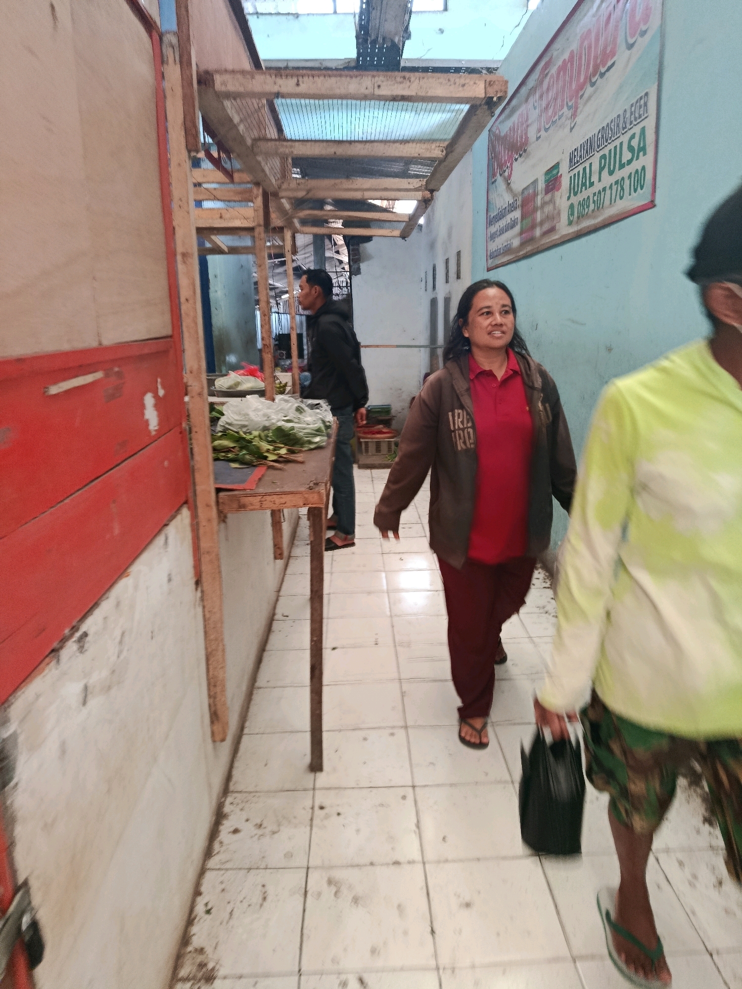 A medium shot captures an outdoor market stall hallway in Bululawang, Indonesia. A woman with dark hair, wearing a red polo shirt and a brown jacket, walks towards the right side of the frame, carrying a black bag. To her right, another person, partially out of focus and wearing a bright green hooded shirt and camouflage shorts, walks away from the camera. In the background, a man in a black jacket stands behind a table laden with produce. The hallway is lined with wooden stalls, one of which displays a banner advertising services and phone numbers. The floor is tiled and appears somewhat dirty.