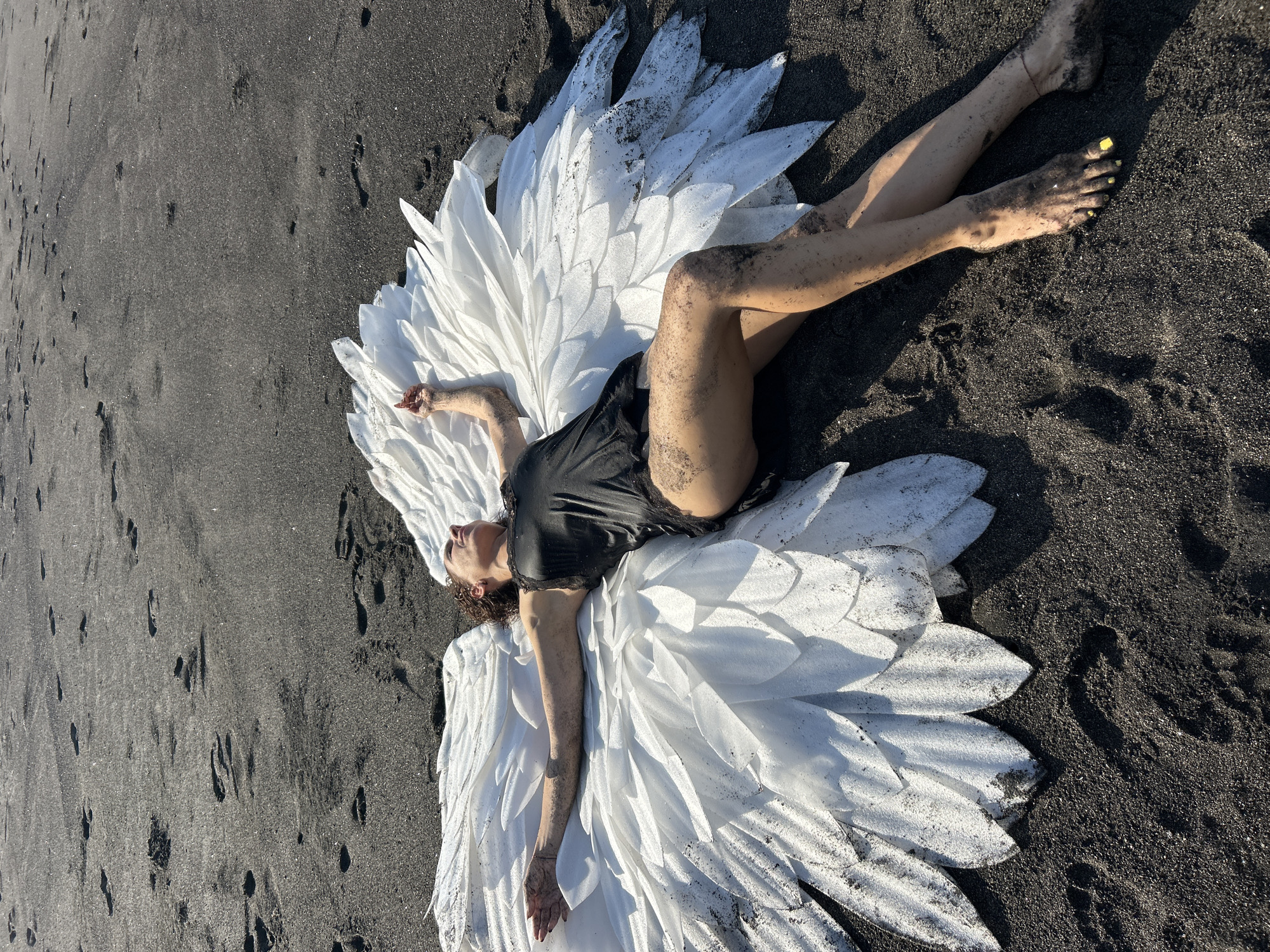 A high-angle shot captures a person, likely female, lying prone on a black sand beach. The individual is positioned centrally, with their body slightly angled. They are wearing a dark, possibly black, top or dress with lace detailing. Spread out beneath and around them is a large, white, feathered or petal-like costume or prop, reminiscent of wings, which creates a striking contrast against the dark sand.
The person's legs and arms are extended, with some black sand visible on their skin and clothing. Yellow nail polish is visible on their toes. Their head is tilted upwards, eyes closed or looking towards the sky, conveying a sense of serenity or repose. The white costume also has traces of black sand on its surface, indicating an interaction with the environment.
The setting is an outdoor beach with fine black sand, characteristic of volcanic regions. The sun is shining brightly, casting distinct shadows from the person and the costume onto the sand, suggesting it is daytime, possibly morning or late afternoon. There are scattered footprints in the sand around the main subject. The overall mood of the image is artistic and dramatic, highlighting the contrast between the white "wings" and the dark sand. No visible text is present in the image.