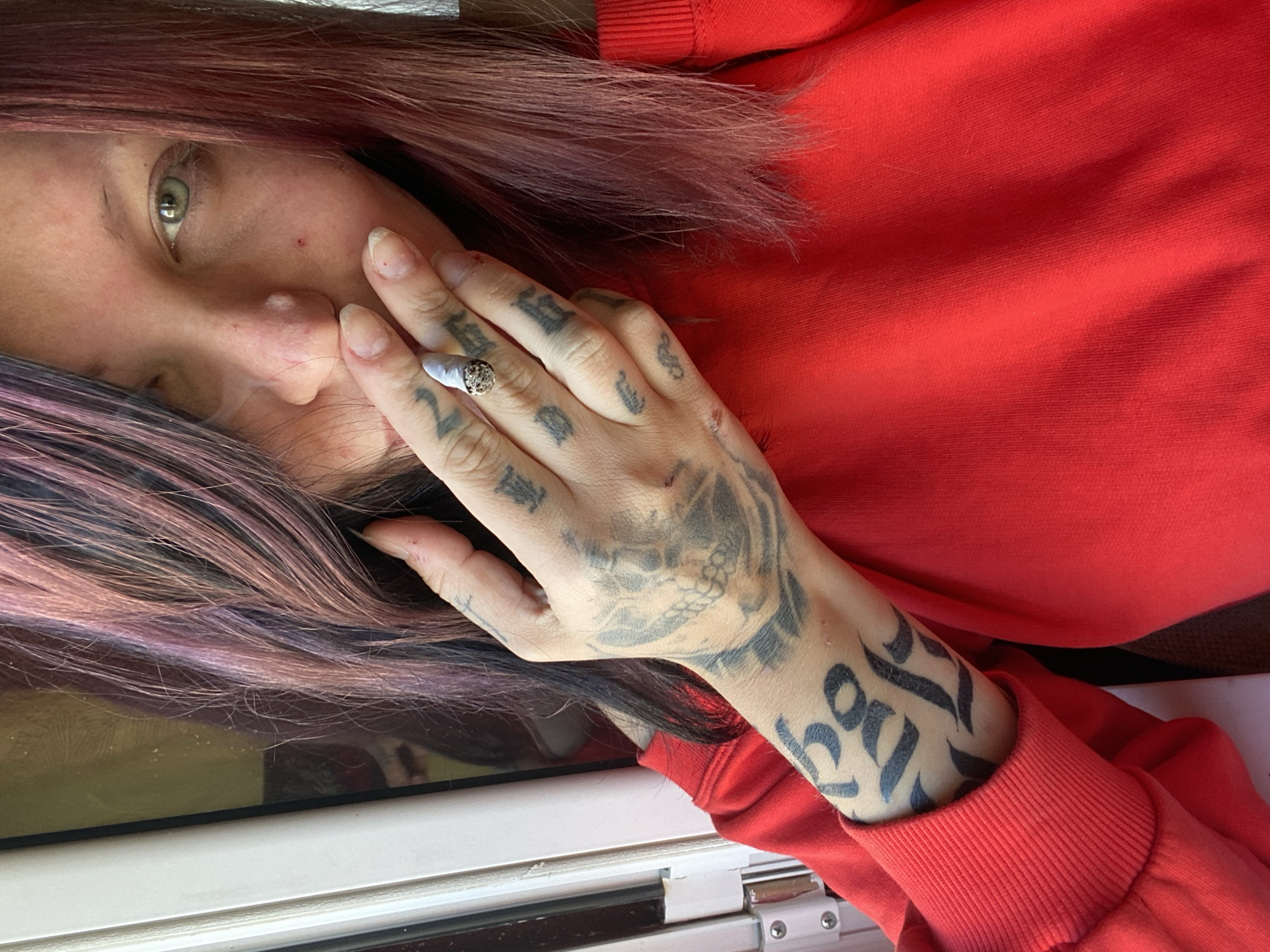 This angled, close-up portrait features a person, likely female, in Kunje, Montenegro, engaged in smoking a lit cigarette or joint. The subject has long, straight hair dyed in distinctive shades of purple, pink, and red, framing one side of their face. They are wearing a bright red long-sleeved top or hoodie, with the ribbed cuff visible on the lower right.
The person's left hand is prominent, heavily adorned with dark tattoos featuring a skull design on the back of the hand and various script-like symbols on the fingers and forearm. A ring is worn on the index finger. The fingernails are long and natural, with some slight reddish marks near the cuticles. The cigarette is held between the index and middle fingers, with a wisp of smoke visible near the nose.
Part of the person's face is visible, showing one green or hazel eye looking towards the left, alongside a portion of their nose and cheek. The expression appears neutral or contemplative. In the bottom left of the image, a white window frame is partially visible, suggesting the scene is either indoors or taken while looking out a window. No discernible text is clearly readable within the tattoos or elsewhere in the image. The scene captures a private, reflective moment.
