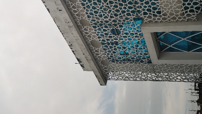The image displays a partial exterior view of a building in Savar, Bangladesh, under an overcast sky. The upper right portion of a multi-story concrete structure is visible, featuring a flat, weathered roofline with dark stains and a slender dark rod protruding vertically. The building's facade is predominantly covered by a light-colored, ornate screen composed of interconnected geometric patterns, resembling an Islamic or Moghul jali design. This screen is positioned in front of blue-tinted glass windows, which reflect a bright, undefined light source. Towards the bottom center, a large rectangular opening, possibly a window or doorway, is framed by a solid gray concrete surround. This opening also contains blue glass, featuring a distinctive white diagonal cross pattern. The overall scene shows no discernible human activity or other prominent subjects beyond the architectural elements and the sky.