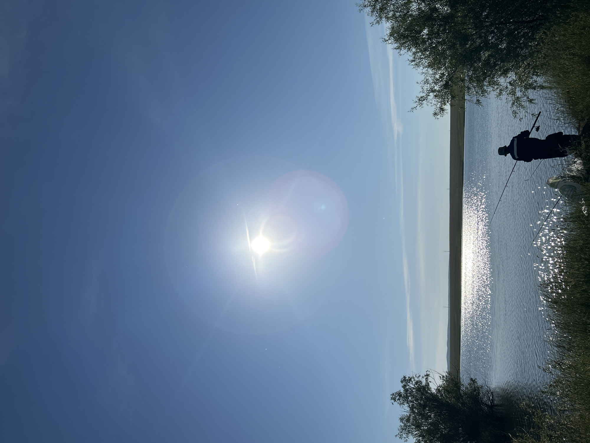 A silhouette of a person is seen fishing on the shore of a lake, with another person in the foreground also fishing. The sun is visible in the bright blue sky, creating a lens flare effect. Trees line the left and right sides of the image, and tall grass grows along the water's edge. In the background, a flat, grassy landscape stretches to the horizon. The scene appears to be in Matrayevo, Russia, likely during a daytime outdoor activity.