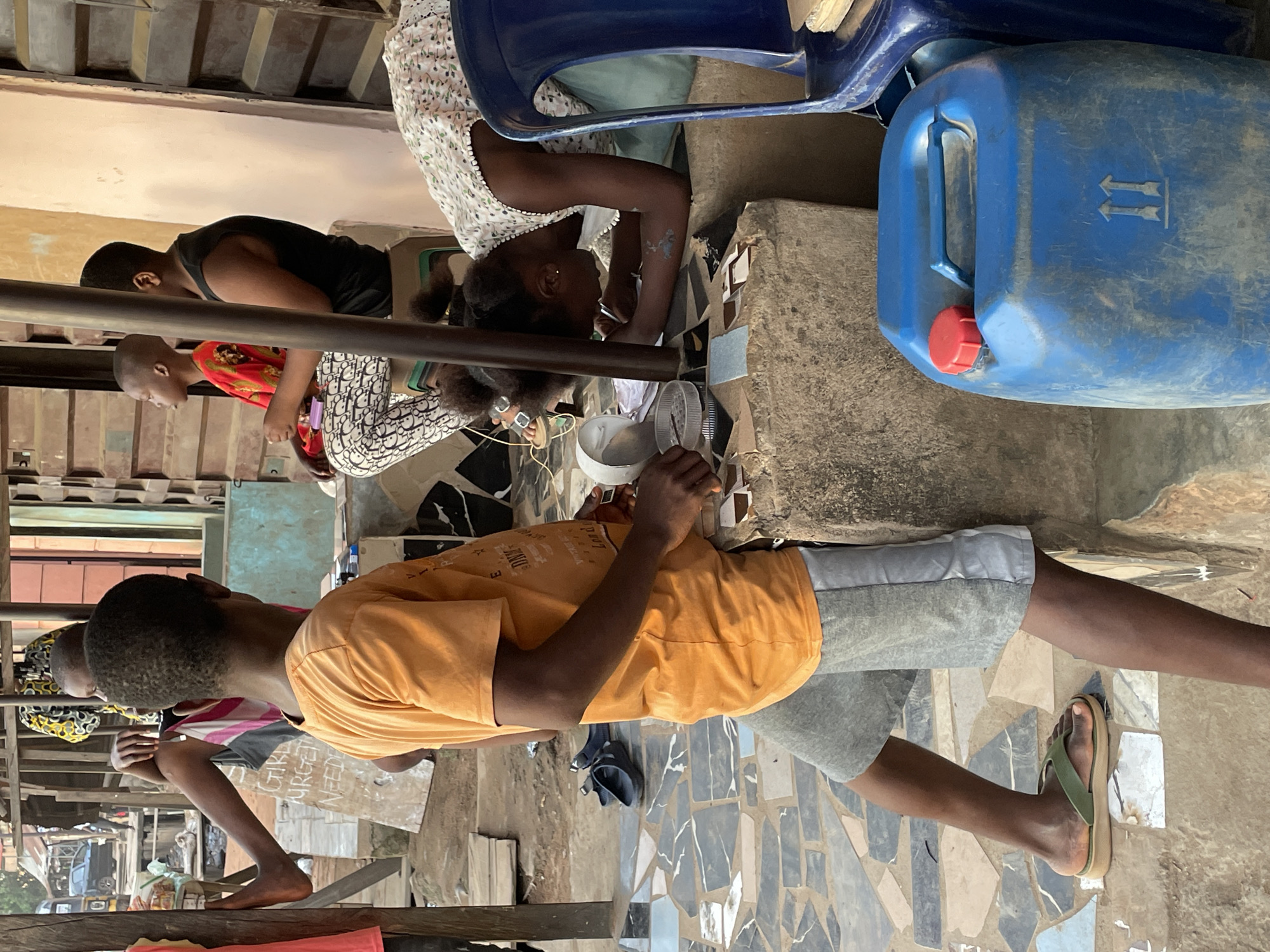 In Nkpor, Nigeria, a group of children are gathered near a wooden structure, possibly a market stall. In the foreground, a boy wearing an orange t-shirt and grey shorts stands, leaning against a concrete block. To his right, a girl in a white floral dress is bent over a collection of small, colorful squares, appearing to be engaged in an activity involving them. She has dark hair pulled back with clips. Behind them, other children are visible. One child, wearing a red shirt and patterned pants, sits with their legs crossed. Another child, in a black tank top and patterned pants, sits to their right. A blue plastic jerrycan with a red cap and a blue handle is prominently placed in the foreground to the right of the frame. The ground is covered with a mosaic of irregularly shaped tiles in shades of gray, white, and blue. The environment suggests an outdoor, possibly commercial, setting.
