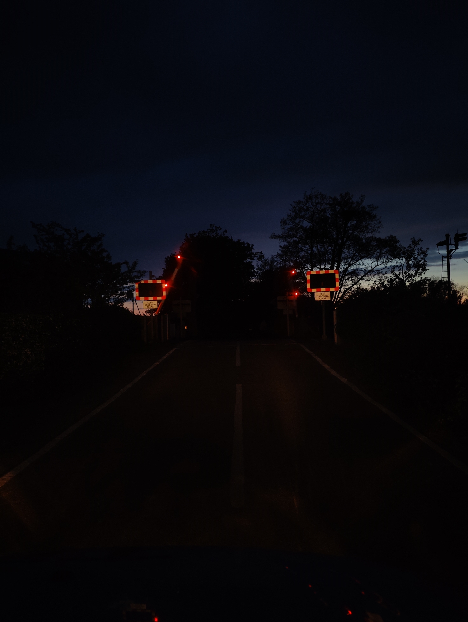 The image shows a road at night in East Suffolk, United Kingdom. Two railroad crossing signs with flashing red lights are visible on either side of the road, indicating a railroad crossing ahead. The signs read "KEEP CROSSING CLEAR." The road is paved with a white dividing line. The surrounding area appears to be wooded with trees silhouetted against the dark sky. There is also what looks like a utility pole with signals on the right side of the image. The scene is dark with the primary illumination coming from the railroad crossing signals.