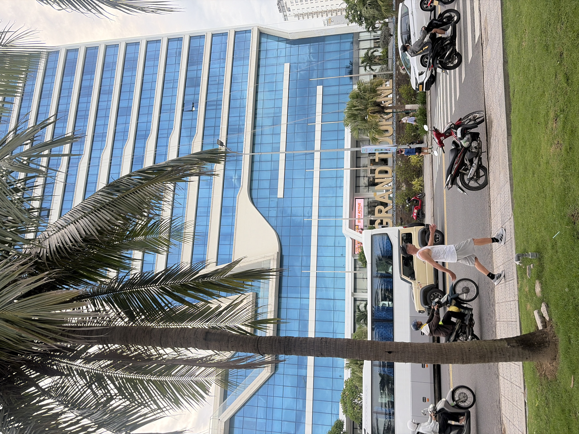 The image captures a street scene in Da Nang, Vietnam, with a large, modern building prominently featured. This building, with its striking blue glass facade and white accents, is identifiable as the Grand Tourane Hotel. In the foreground, a man in a white tank top and grey shorts is walking across a pedestrian crosswalk, appearing to be mid-stride. He is wearing athletic shoes with grey and white details and black socks.
Several vehicles are present on the road. To the left, a white tour bus with tinted windows is parked or moving slowly. A woman on a scooter, wearing a white helmet and jacket, is visible on the left side of the bus. Further down the road, a beige SUV-like vehicle is positioned behind the bus. A man on a dark-colored motorcycle, wearing a brown jacket and blue helmet, is riding towards the right.
On the right side of the crosswalk, a white SUV is visible, and a person on a black motorcycle, wearing a grey hooded jacket and a black helmet, is crossing the street. A red scooter is parked near the curb on the left side of the image.
The environment is urban, with palm trees lining the street and a well-maintained grassy area with a few scattered rocks and a small pipe visible in the foreground. The sky is overcast, suggesting a cloudy day. The overall impression is one of bustling city life with traffic and pedestrian activity.