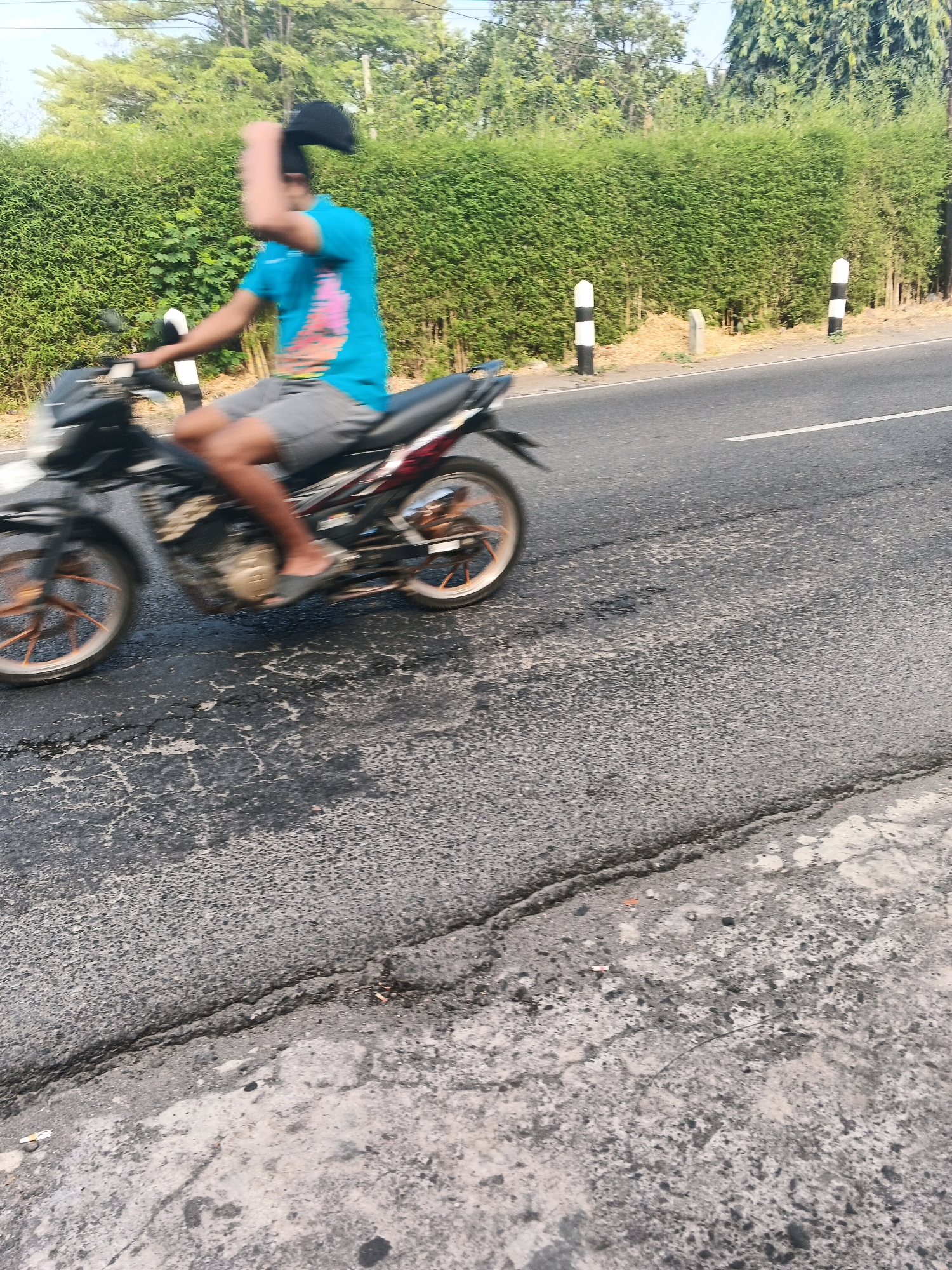 A person wearing a blue t-shirt and gray shorts rides a black motorcycle on a cracked asphalt road in Ungaran, Indonesia. The person is holding a black cap in their right hand, which is blurred due to motion. The motorcycle has orange rims and a black seat. A dense green hedge lines the right side of the road, with two white and black striped posts visible along it. The sky is overcast, and trees are visible behind the hedge. The image appears to have been taken during the day.