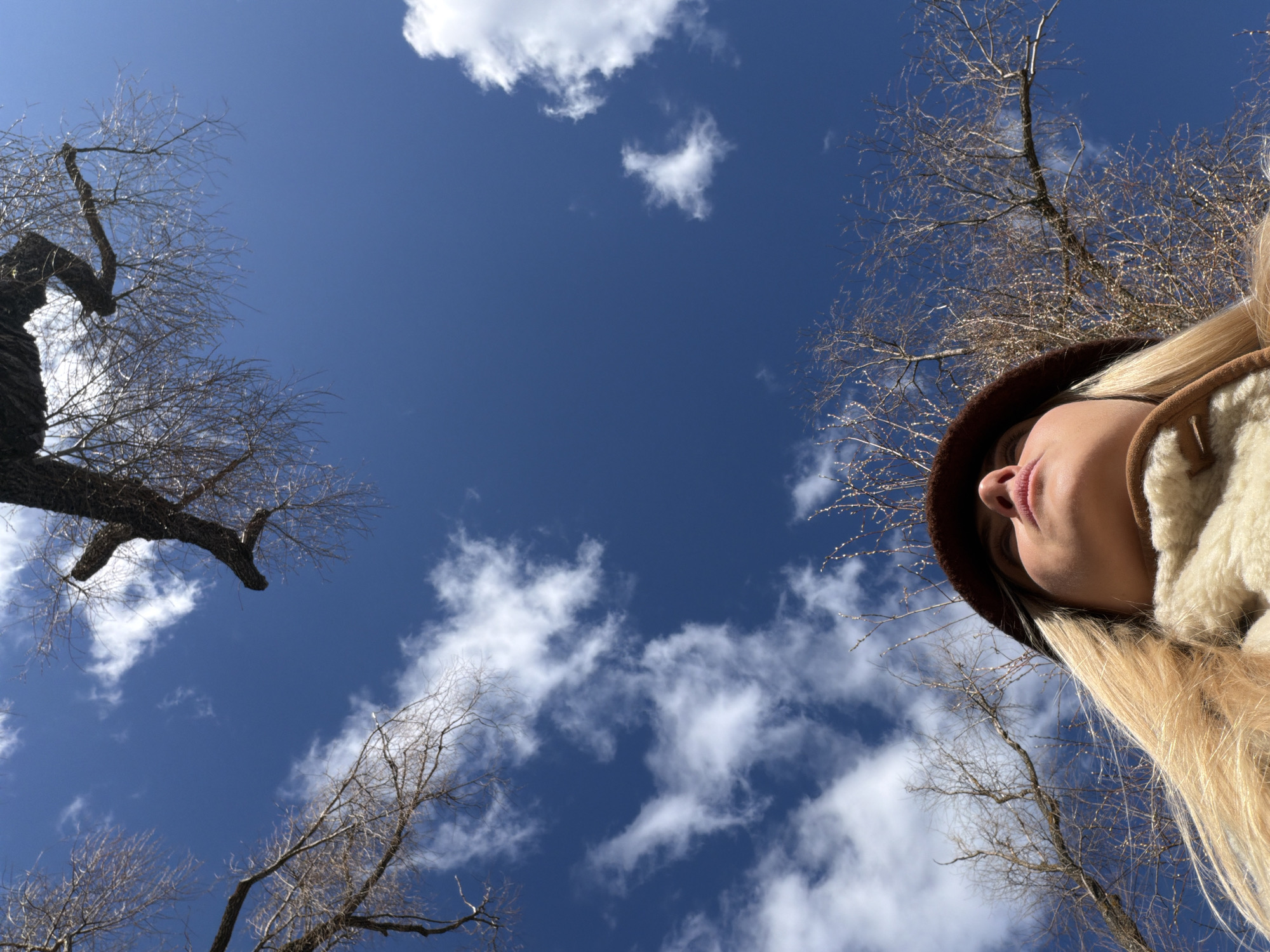 A low-angle shot captures a person partially framed against a blue sky with scattered white clouds and bare trees. The person, located in the lower right of the frame, has blonde hair, a dark brown hat, and appears to be wearing a light-colored jacket. Only the face and upper torso are visible. Bare tree branches extend into the sky from the left and right sides. The location appears to be outside, potentially in a park or wooded area. Specific identification of the location as Votkinsk, Russia is not possible based solely on the provided image.