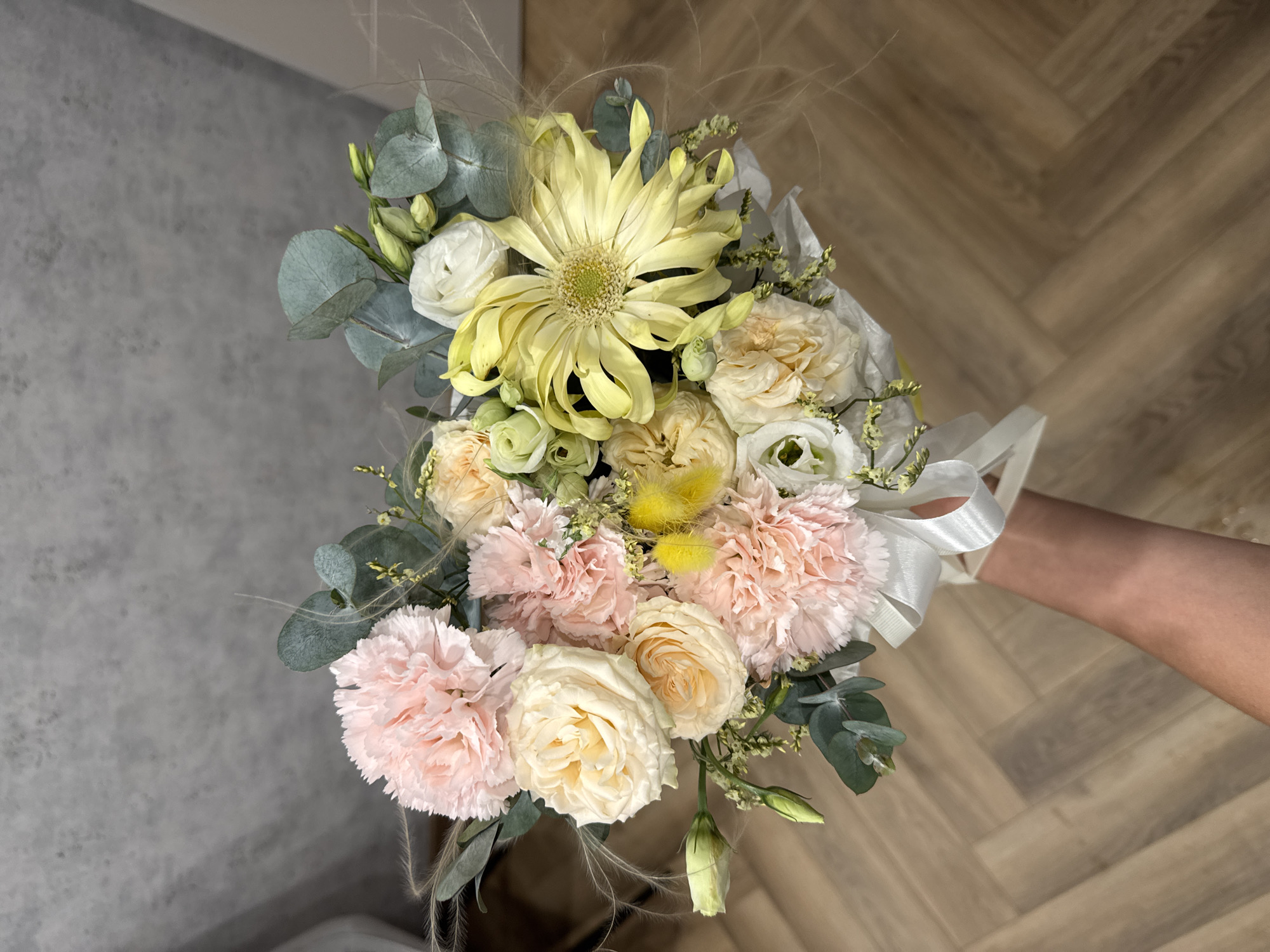 A bouquet of flowers is held up in front of a gray and light brown herringbone patterned background. The bouquet contains pink carnations, light yellow roses, a large yellow flower with narrow petals, white flowers, and greenery including eucalyptus leaves. A white ribbon is tied around the stems of the bouquet. A person's arm is visible holding the bouquet. The location is not specified, and no city is mentioned.