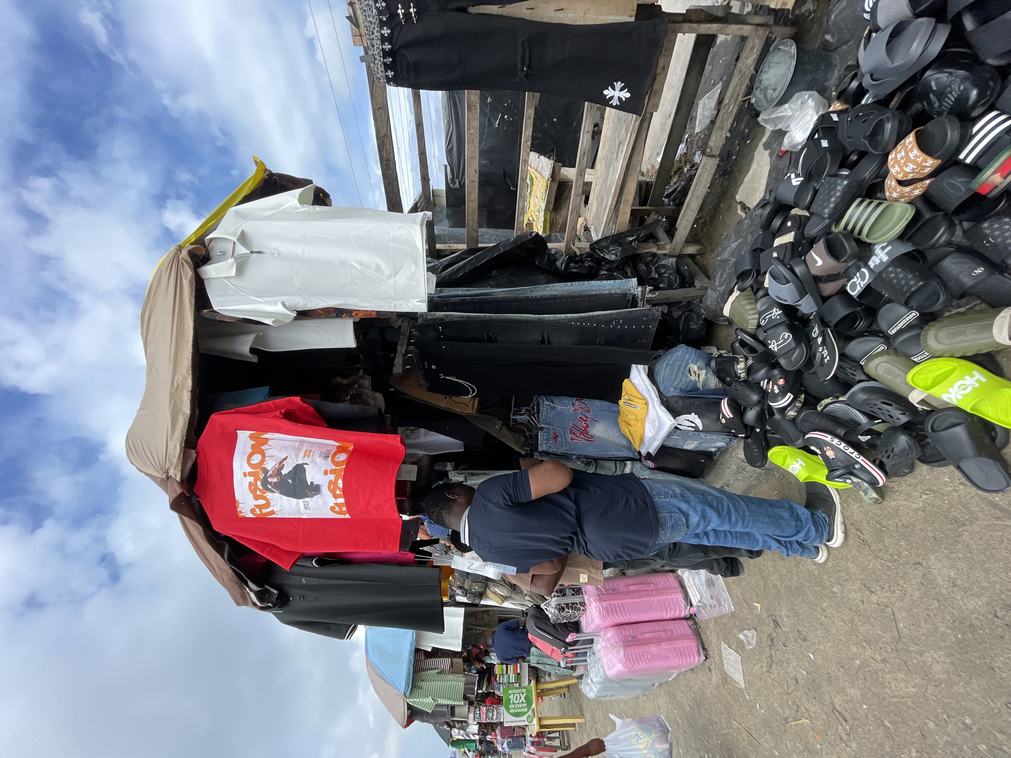This is a street market scene in Lekki, Nigeria, on a cloudy day. A stall displays various clothing items, including a red t-shirt with the word "fusion" and an image of a person, a black polo shirt, and a white polo shirt with a zipper. Hanging below are black pants and denim jeans, some with rips. A large pile of assorted flip-flops and slides is visible on the ground in front of the stall. Two men are present; one is standing by the stall, looking at the merchandise, and another is partially visible further down the market. Pink suitcases are stacked near the stall. The environment appears to be an outdoor market with makeshift structures and goods laid out for sale.