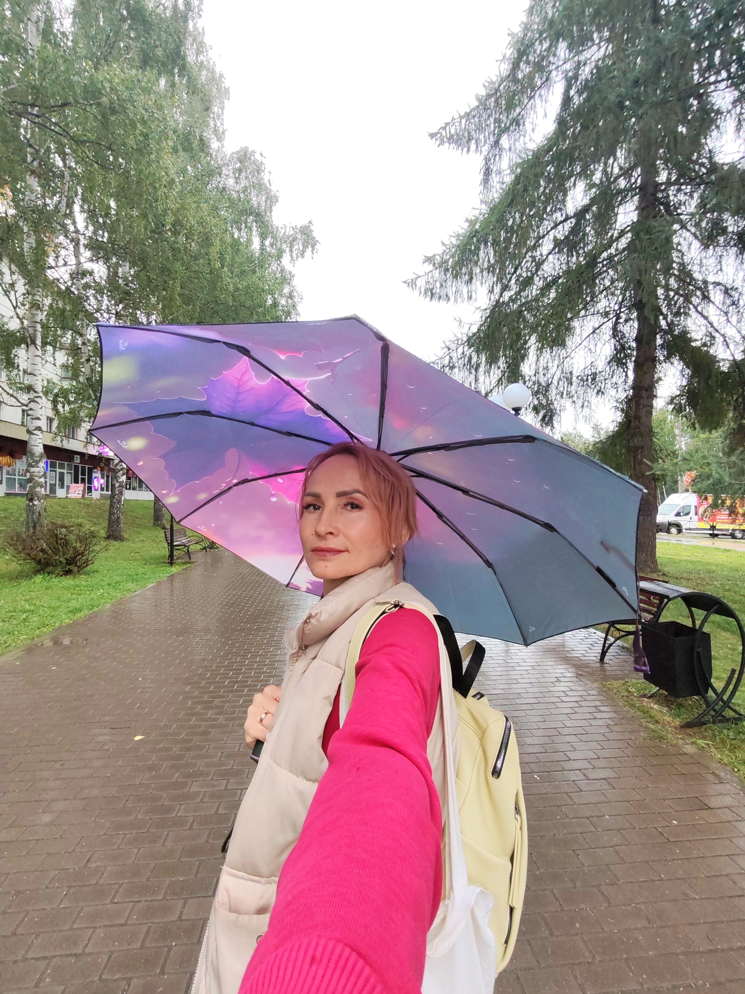 A woman with pink hair stands on a wet, brick-paved sidewalk holding a large umbrella patterned with purple and pink leaves. She is wearing a cream-colored vest over a bright pink sweater and a yellow backpack. The umbrella is open, obscuring most of her upper body and the sky. Trees with green leaves line the sidewalk, and in the background, there are buildings and a white van. The scene appears to be an outdoor urban environment, possibly a park or street in Chaikovsky, Russia, on a cloudy or rainy day, indicated by the wet pavement and umbrella.