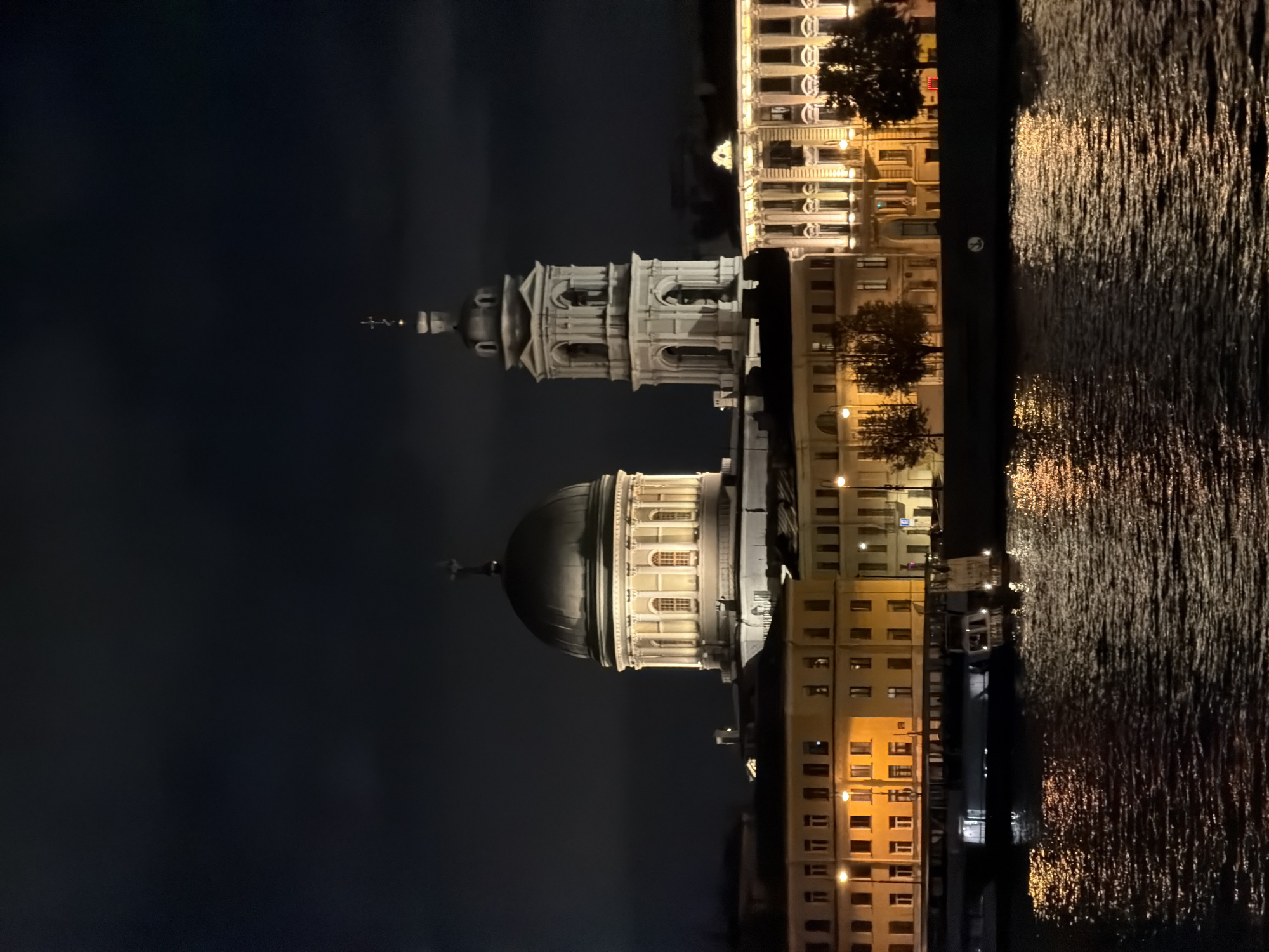 The image captures a nighttime view of the Saint Isaac's Cathedral in Saint Petersburg, Russia, reflected in the dark waters of the Neva River. The cathedral's grand dome and bell tower are illuminated, creating a majestic silhouette against the night sky. Several boats are docked along the riverbank, their lights twinkling on the water. The buildings lining the river are also lit, adding to the vibrant urban scenery. The overall atmosphere is one of serene beauty and architectural grandeur.