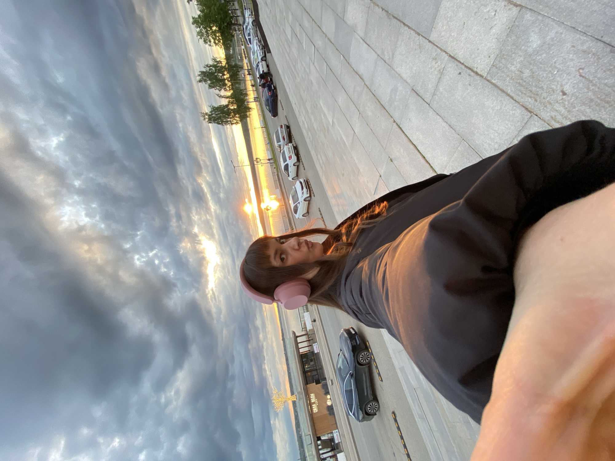A selfie of a young woman wearing pink headphones, with a view of the Kama River waterfront in Votkinsk, Russia, during a sunset or sunrise. The woman is wearing a dark jacket. Behind her, a row of parked cars lines the waterfront promenade. The sky is cloudy, with the sun partially visible, reflecting on the water. Buildings are visible in the background. The foreground consists of a textured concrete surface.