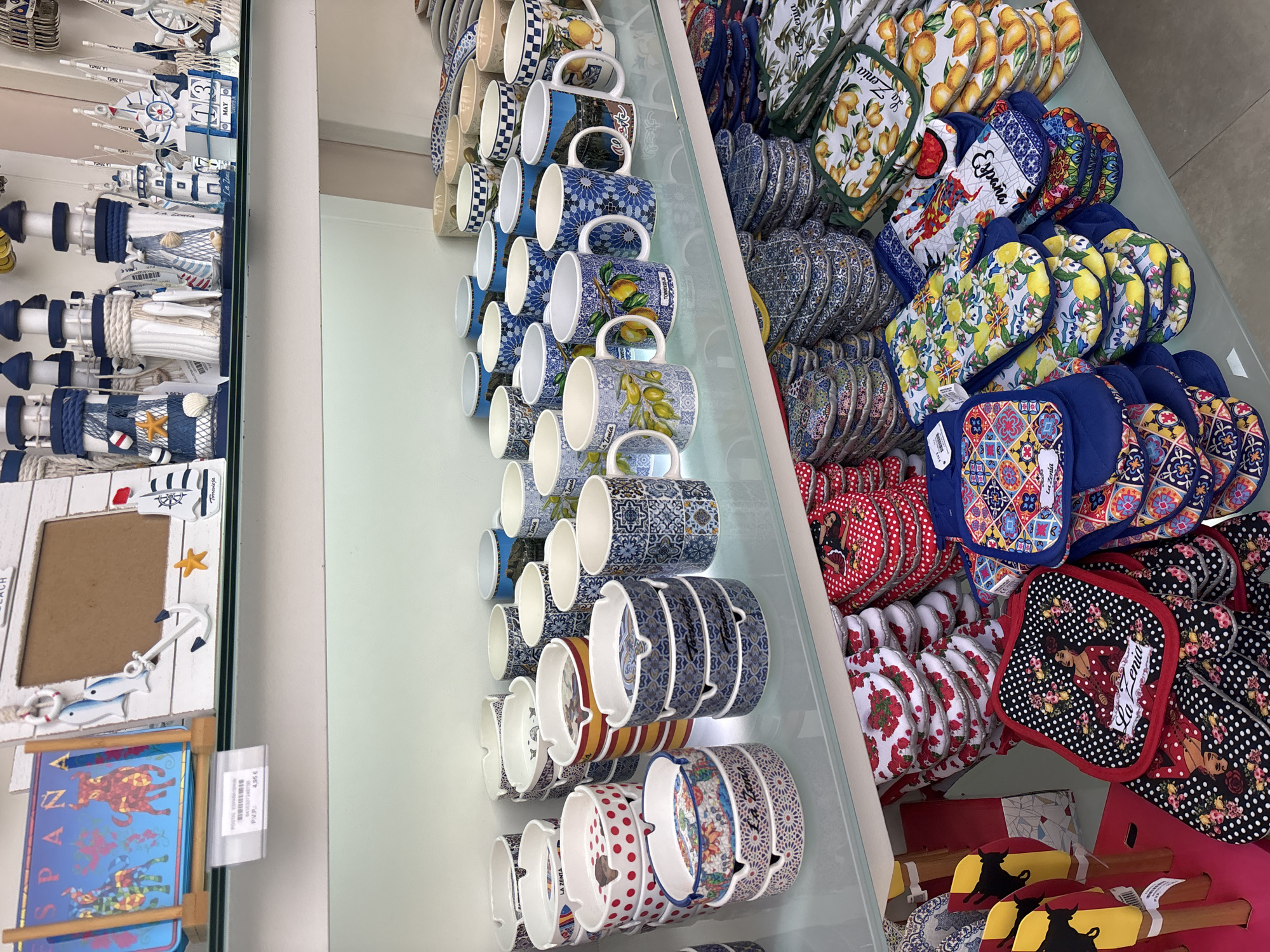 The image displays a retail display of souvenirs and kitchenware items on multiple shelves. The top shelf features nautical-themed decorative items, including lighthouse figurines and miniature sailboats, along with a framed photo holder. A price tag indicates "ESPAÑA" and "P.V.P. 4,95 €". The middle shelf showcases a variety of ceramic mugs with diverse designs, some featuring scenic landscapes, citrus fruits, and geometric patterns. Below this, several stacks of ashtrays are arranged, some with polka dot patterns and others with mosaic-like designs. The bottom shelf is filled with oven mitts and potholders, many of which are patterned with floral motifs, lemons, and Spanish-themed imagery, including one with a flamenco dancer. The items suggest a souvenir shop, likely located in a tourist area of Orihuela, Spain, given the "ESPAÑA" label and the common souvenir themes. There are no people visible in the image, and the environment appears to be an indoor retail setting.