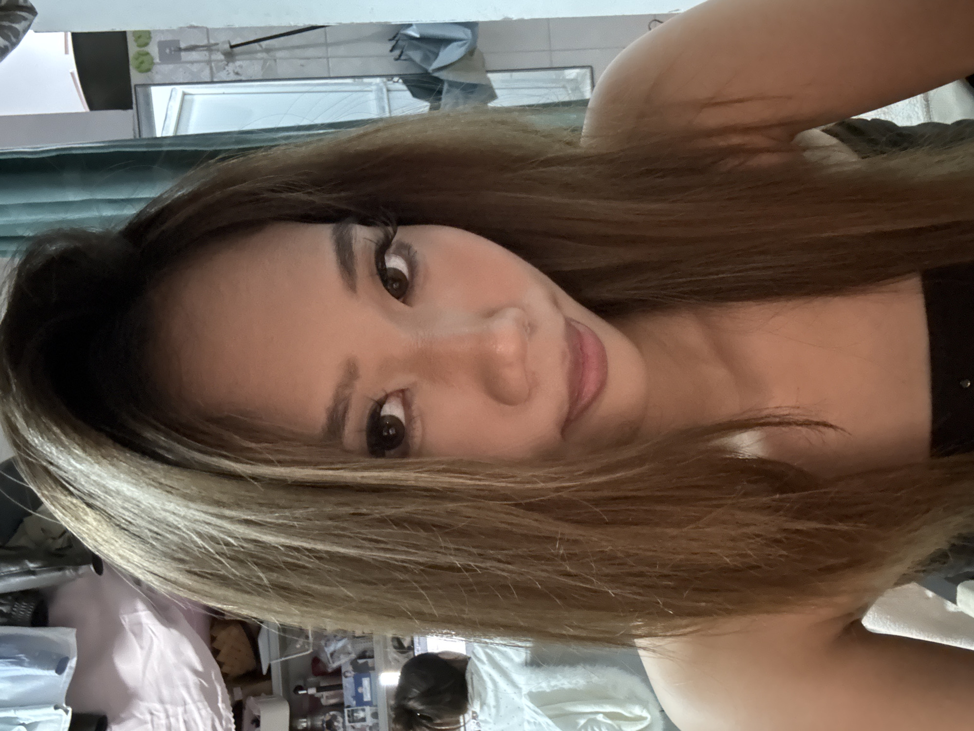 A selfie of a woman with long, light brown hair is centered in the frame. Her face is tilted slightly to the right, and she is looking at the camera. The background is a blurry interior scene. A bathroom with a mirror and a shower curtain can be seen behind her. At the bottom of the frame, there is a cluttered space that appears to be a desk or table with various items. The location is in the Nan'an District, China.