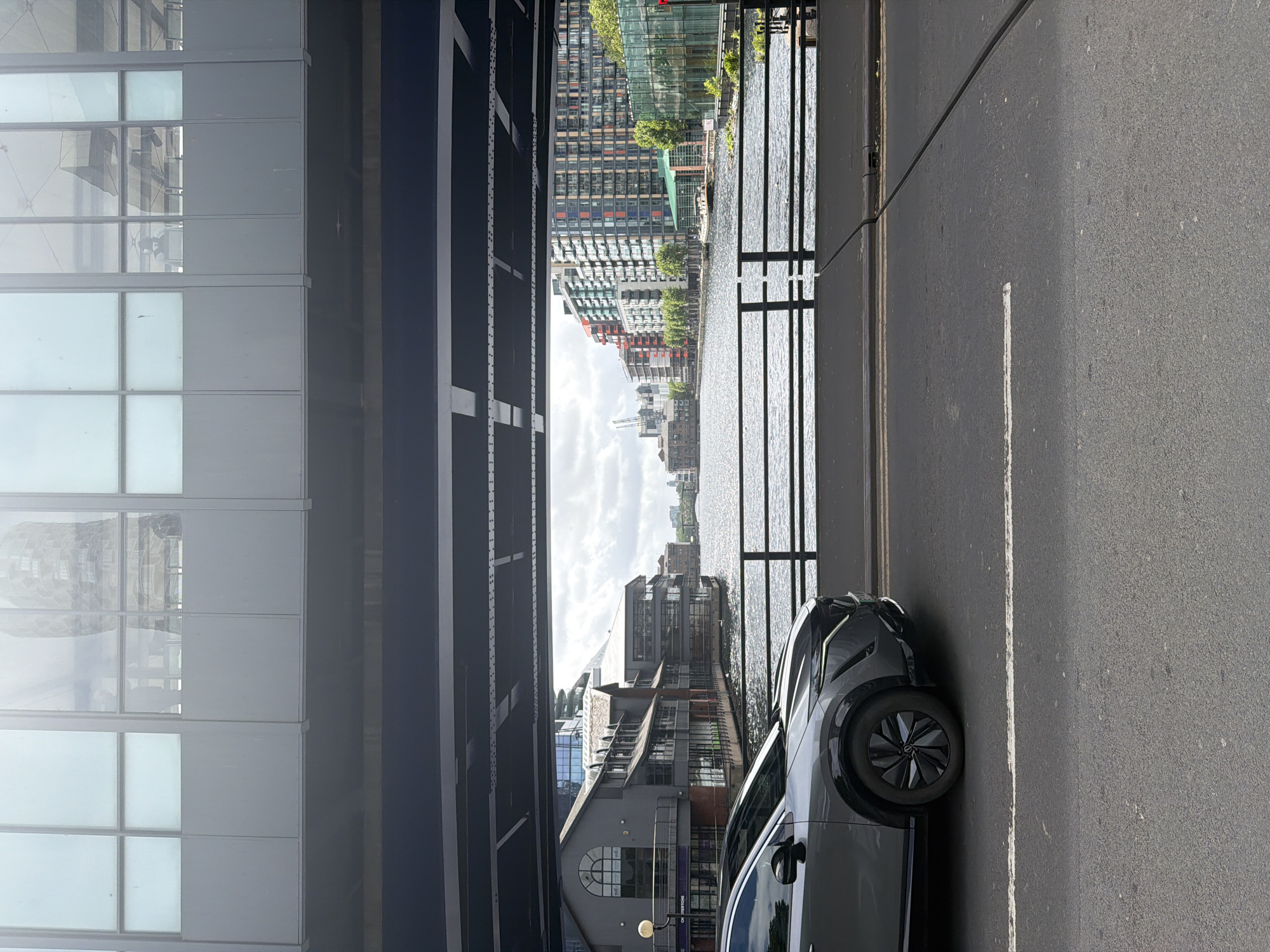 The photograph presents an urban landscape, likely in London, United Kingdom, characterized by modern architecture and infrastructure. The scene is partially framed by the structure of a bridge or elevated roadway, composed of dark steel beams and supports. To the left, a contemporary building with large, gridded windows dominates the view. In the central background, a body of water reflects the sky and surrounding buildings. Along the riverbank are densely packed buildings of varied architectural styles, with residential or commercial units having multiple windows and some greenery interspersed.
On the right side of the frame, a dark-colored car is parked on the pavement beside a metallic fence. The pavement, marked with a road line, is visible in the foreground.