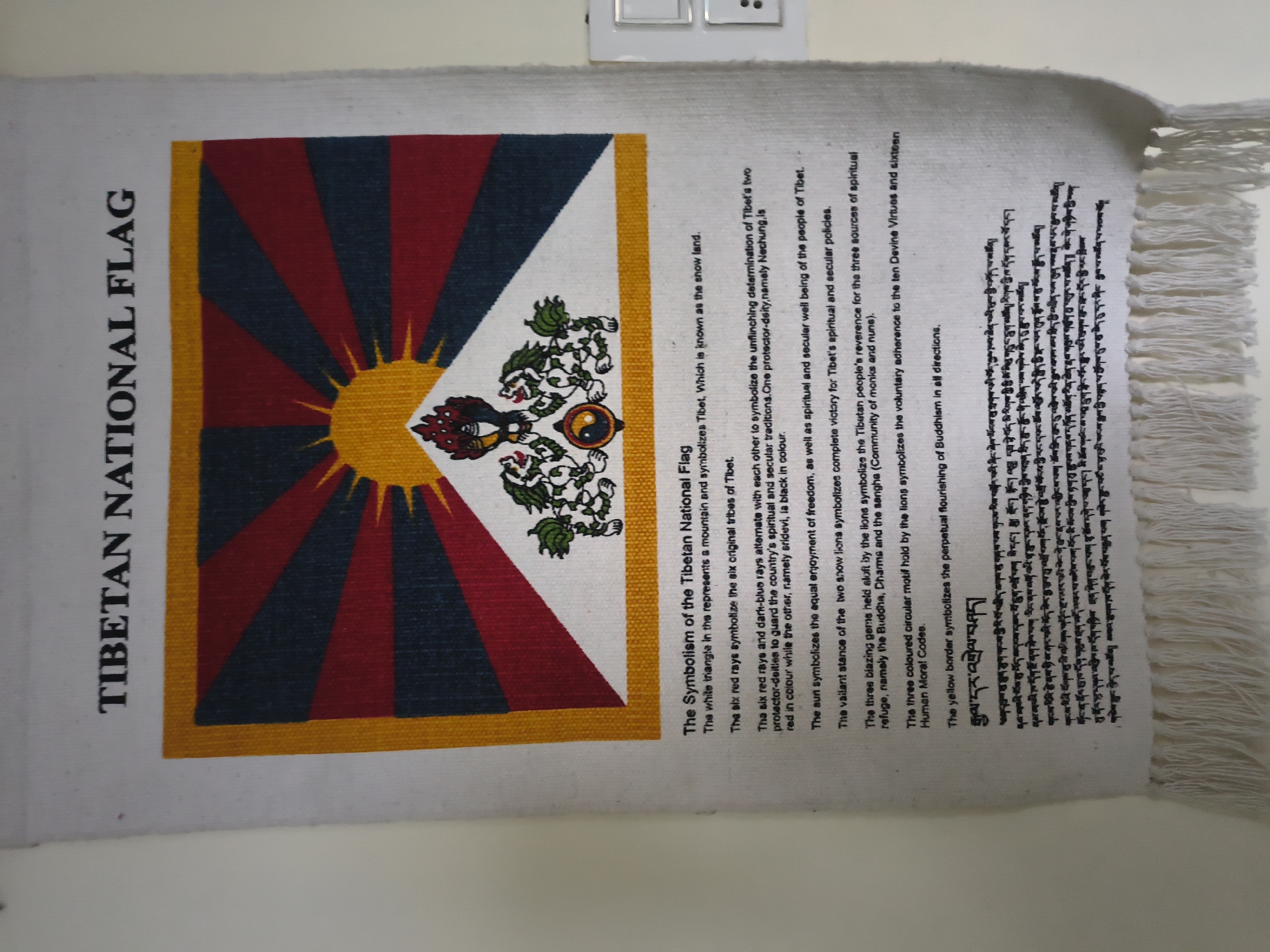 The image shows a tapestry of the Tibetan National Flag hanging on a wall. The tapestry is white with a prominent depiction of the flag, which features a sunburst, mountains, and two snow lions holding a flaming jewel. Below the flag, there is text describing the symbolism of the flag, written in English and Tibetan script. The text is presented in two columns, with the English explanation on the left and the Tibetan script on the right. The tapestry has a frayed, fringed edge at the bottom. The wall appears to be a neutral color, possibly white or light grey. There is a light switch plate visible on the right side of the image. The image is taken from a medium distance, capturing most of the tapestry and a portion of the surrounding wall. There are no people visible in the image. The scene appears to be indoors, possibly in a home or a cultural institution. There is no information to indicate the location is in Haini, India; this information is not visually supported by the image.