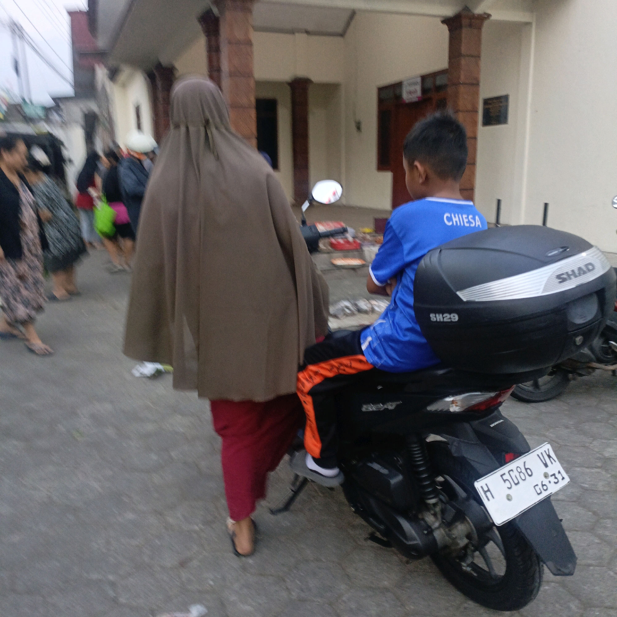 In Salatiga, Indonesia, an outdoor, street-level scene depicts several individuals and a motorcycle. In the foreground, a person wearing a brown hijab and red pants is walking away from the camera, obscuring much of the lower portion of a motorcycle. Seated on the motorcycle's rear is a young boy wearing a blue t-shirt with the white text "CHIESA" and orange and black pants. The motorcycle is black with a prominent black storage box on the back labeled "SHAD" and "SH29". The license plate of the motorcycle reads "H 5086 VK" with a date stamp "06.31". To the left of the hijab-clad person, other individuals are visible. One woman in a patterned dress walks away from the camera, and behind her, another person in a grey and black patterned skirt and a dark top is also walking away. Further left, a person in a black top and jeans stands with their back to the camera. In the background, a building with brick columns and cream-colored walls is visible. The ground is paved.