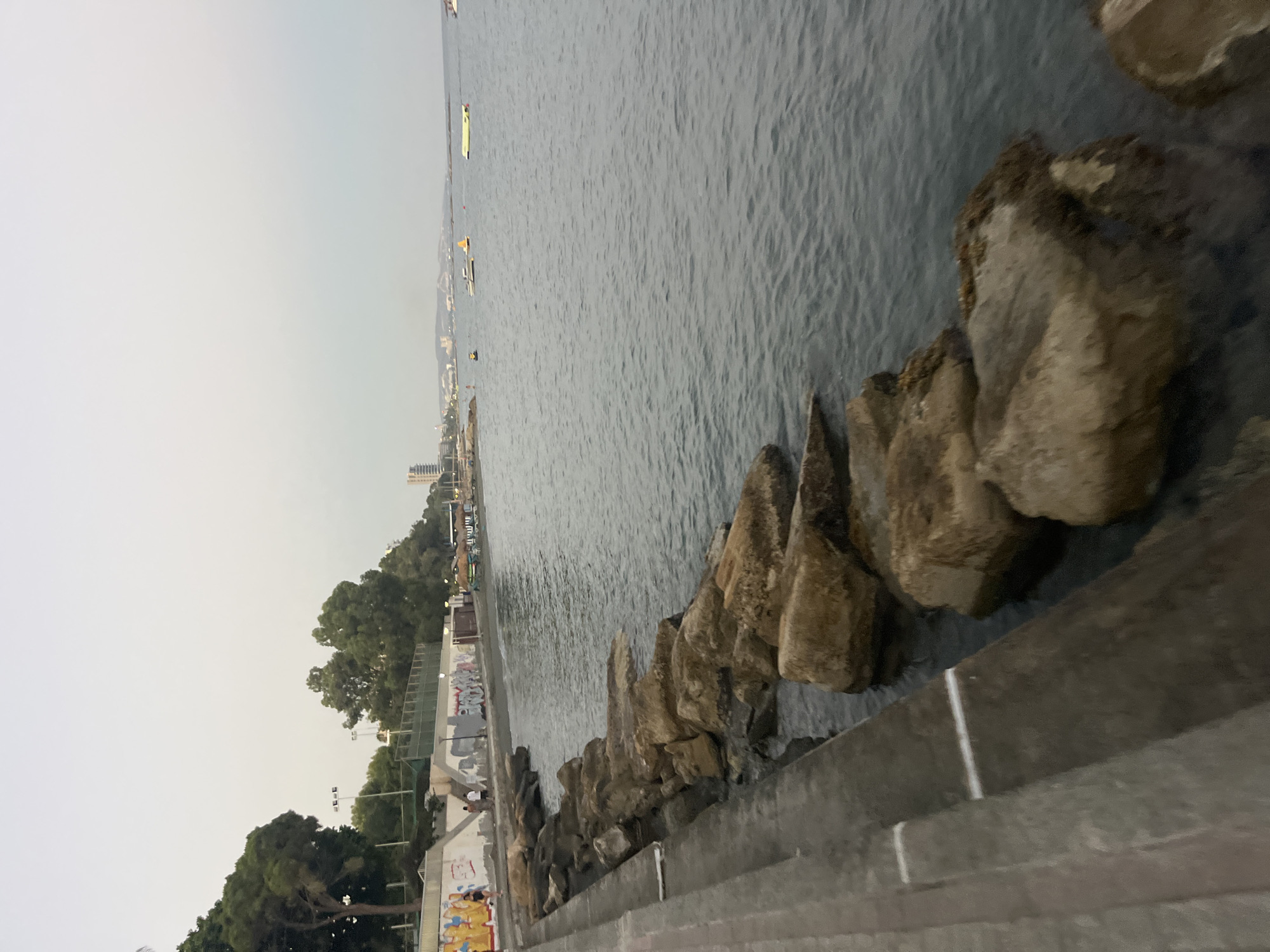 The image depicts a section of the coastline in Agios Tychona Community, Cyprus, likely during the late afternoon or early evening due to the muted lighting. In the foreground, a concrete walkway runs along the left side of the frame, with several large, light-brown rocks lining the edge of the water. The sea is calm with gentle ripples.
Beyond the walkway and rocks, a beach area is visible, backed by a low retaining wall adorned with graffiti. Several trees with dark green foliage are planted behind the wall, and a few streetlights are present. In the distance, a few small boats and what appear to be yellow buoys are scattered on the water. The background reveals a hazy skyline with buildings, indicating a developed coastal area. A few individuals can be seen walking along the beach and the walkway. The overall atmosphere is serene and coastal.