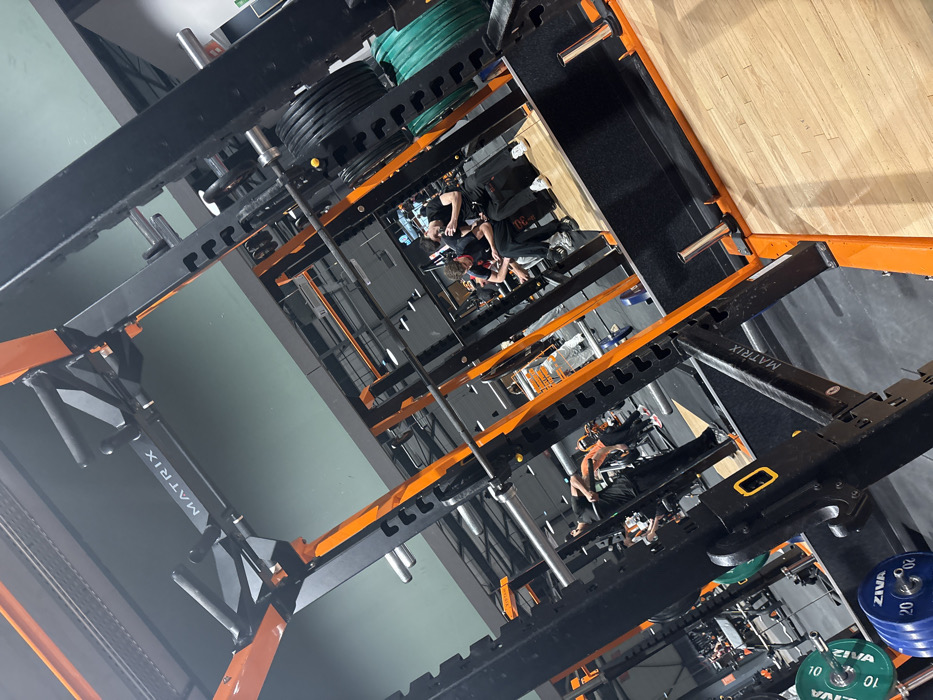 A tilted view showcases a gym interior with several squat racks featuring black and orange frames, along with weight plates. People are visible in the racks and on the floor. The racks are labeled "MATRIX". The floor appears to be a mix of wood and darker material. The image has an unusual angle that creates a sense of depth and complexity.