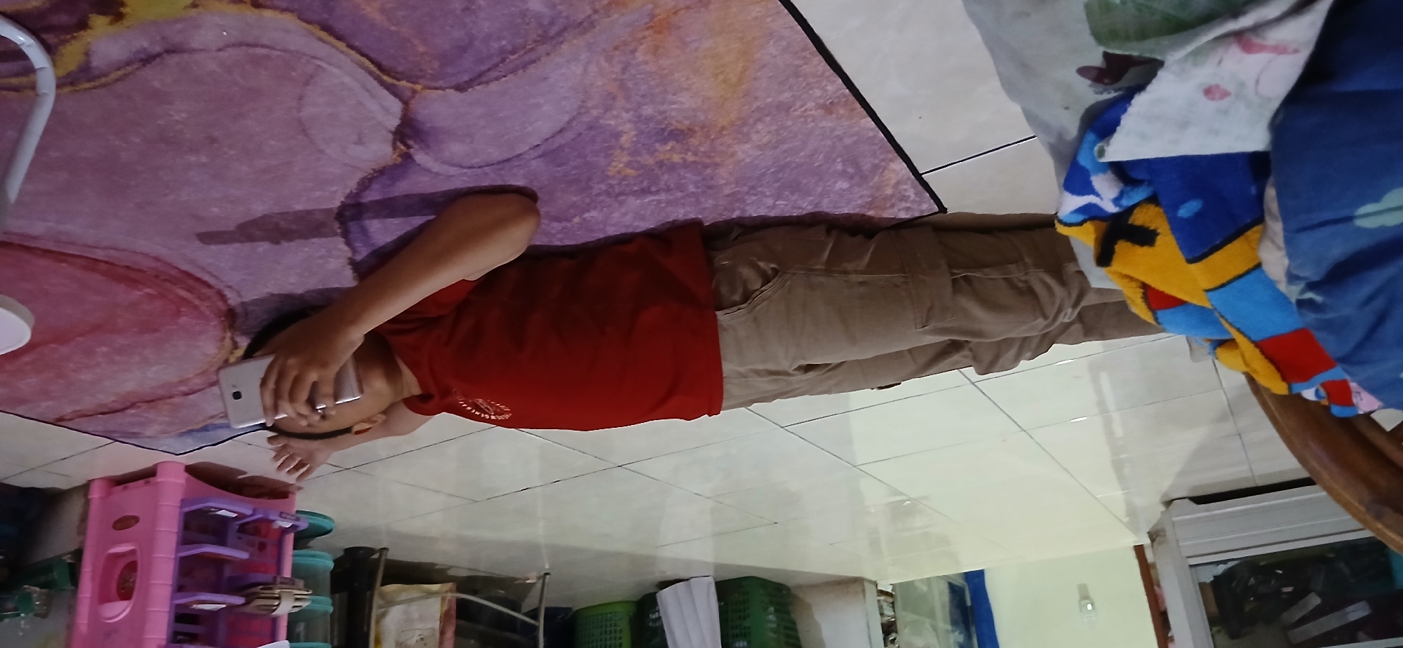 A young person is lying on their back on a floor covered by a purple and pink rug. They are wearing a red t-shirt and tan pants. The person is holding a white smartphone up to their right ear with their right hand, as if making a call or listening to something. Their left arm is resting above their head. A colorful blanket with blue, yellow, and red geometric patterns is bunched up near their legs. The floor is tiled with large, light-colored square tiles. In the background, there is a white cabinet with glass doors, a green plastic basket, and a pink plastic toy shelf with purple shelves. The scene appears to be indoors, possibly in a residential setting in Samarinda, Indonesia.