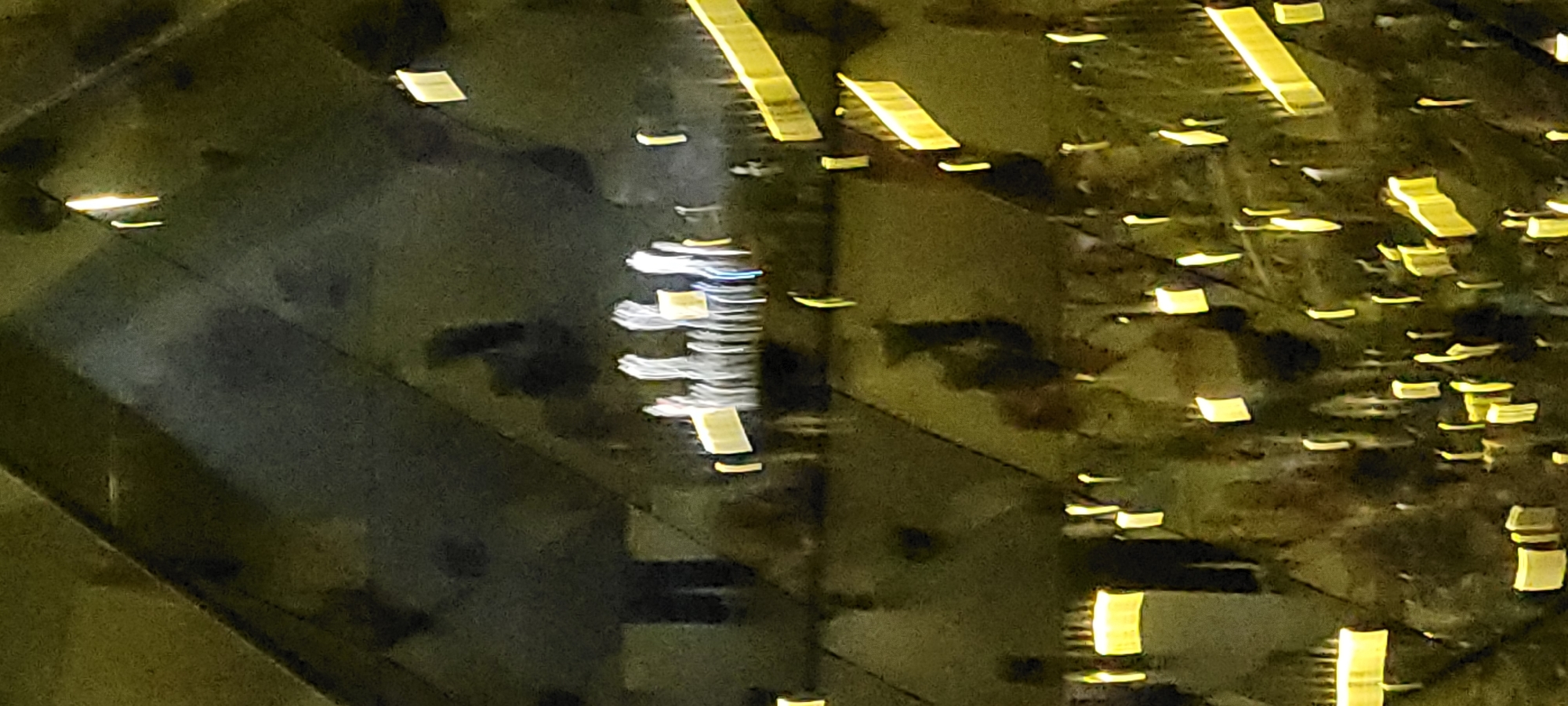 The image depicts an interior space with geometric architectural features, likely a modern building or art installation. The lighting is dim and artificial, with numerous rectangular and elongated yellow lights scattered throughout the scene, creating reflections. In the upper portion of the image, a group of figures, possibly people, are visible as blurred, elongated white shapes with some yellow elements. They appear to be standing in a line or cluster. The overall impression is one of abstract light and shadow, with a sense of depth created by the reflections. The location is identified as Wattens, Austria, which is known for its Swarovski Crystal Worlds, a popular tourist attraction featuring crystal art installations and architectural wonders. Given the abstract and reflective nature of the image, it is plausible that this photograph was taken within the Swarovski Crystal Worlds.