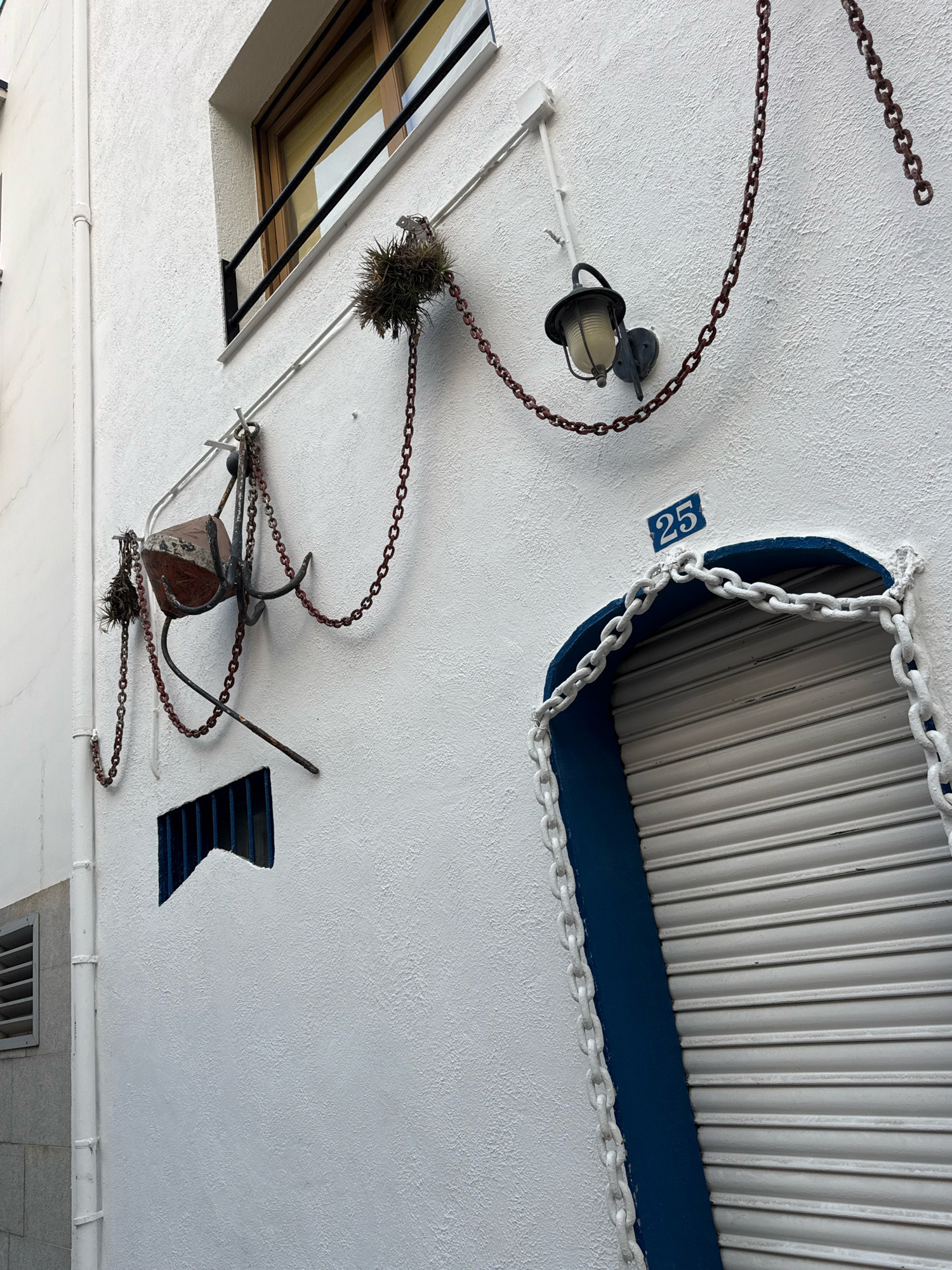 The image shows a white stucco wall in Blanes, Spain, with several nautical-themed decorations. A window with dark metal bars is visible in the upper left. Below the window, a rusty chain drapes across the wall, attached to a small anchor and a buoy-like object. Further to the right, a wall-mounted lantern hangs. Dominating the right side of the image is an arched doorway, painted navy blue, with a closed, white metal rolling shutter. A thick, white chain encircles the opening of the arched doorway. The number "25" is displayed on a blue tile above the doorway.