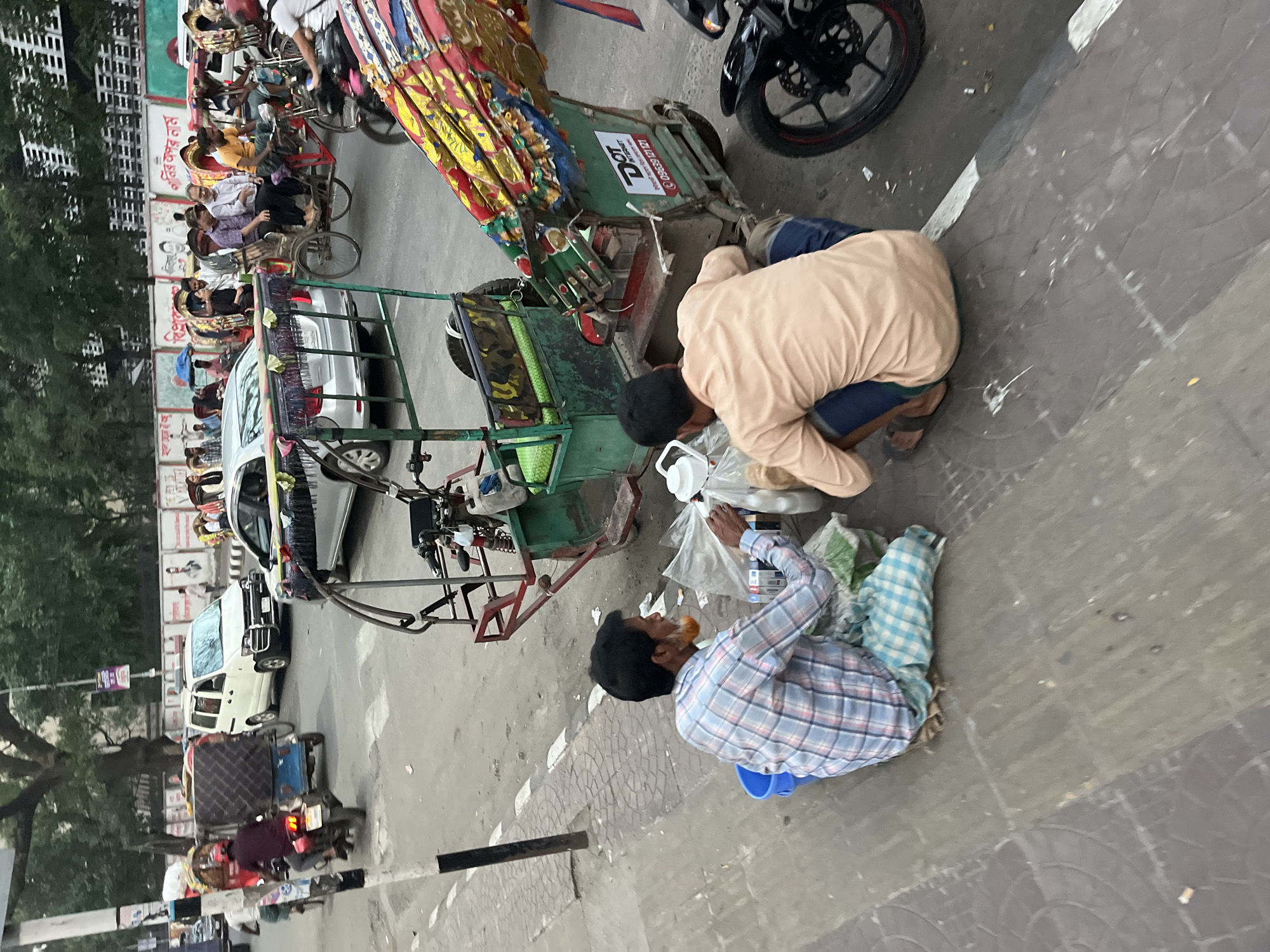 The image shows a street scene in Dhaka, Bangladesh. Two men are squatting near a rickshaw on a paved street. One man is wearing a light brown shirt and dark shorts, while the other is wearing a plaid shirt. Both appear to be examining or sorting through items in plastic bags. In the background, there are several rickshaws, cars, and motorcycles, indicating a busy urban environment. Trees line the street, and a tall building is visible in the distance.