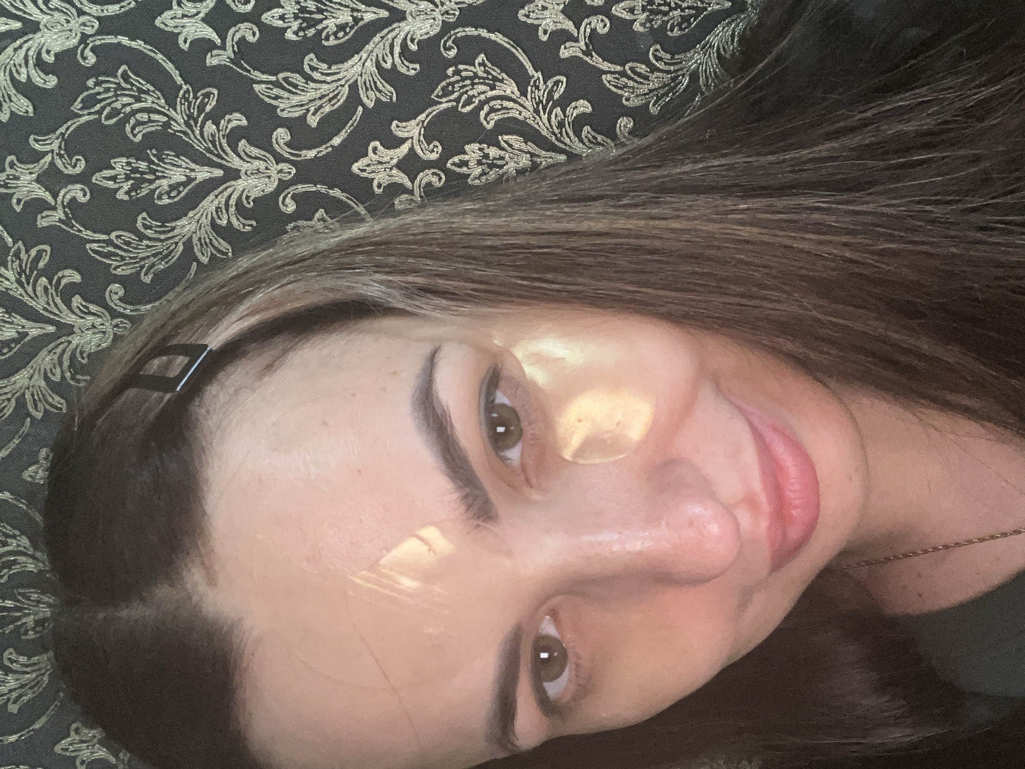 A woman is captured in a close-up, angled shot, appearing relaxed and content. Her head is tilted, suggesting she is lying down or leaning against a patterned surface. She has long, straight, dark brown hair, with a small, black, geometric hair clip visible on the left side of her head. Her face is towards the camera, revealing brown eyes, dark eyebrows, and a soft, faint smile on her pink lips.
Notable details include two bright, reflective patches of light on her face: one across her forehead and the bridge of her nose, and another on her right cheekbone, possibly from a window or light source. She is wearing a thin, gold chain necklace, faintly visible around her neck.
The background consists of a dark fabric or surface adorned with an intricate, ornate gold or silver floral and leaf pattern, hinting at an indoor setting such as a living room or bedroom, possibly a decorative pillow, sofa, or headboard. The overall mood is calm and introspective. No text is visible.