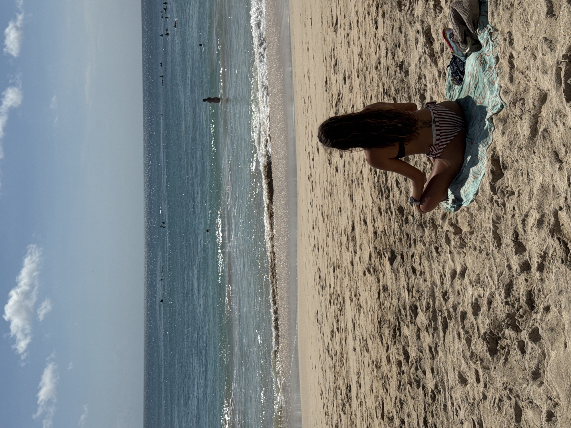 The image depicts a sunny beach scene in Kuta Selatan, Indonesia. The beach extends along the right side of the frame, revealing a textured tan sand surface marked with various patterns and depressions. A person in a striped bikini sits on a blue and green towel, partially obscured by clothing and other items placed nearby. The turquoise water of the ocean occupies the left side, reflecting sunlight and displaying gentle waves reaching the shore. In the distance, several individuals are visible in the water, enjoying swimming and other water activities. Above the ocean, a blue sky with scattered white clouds completes the serene backdrop.