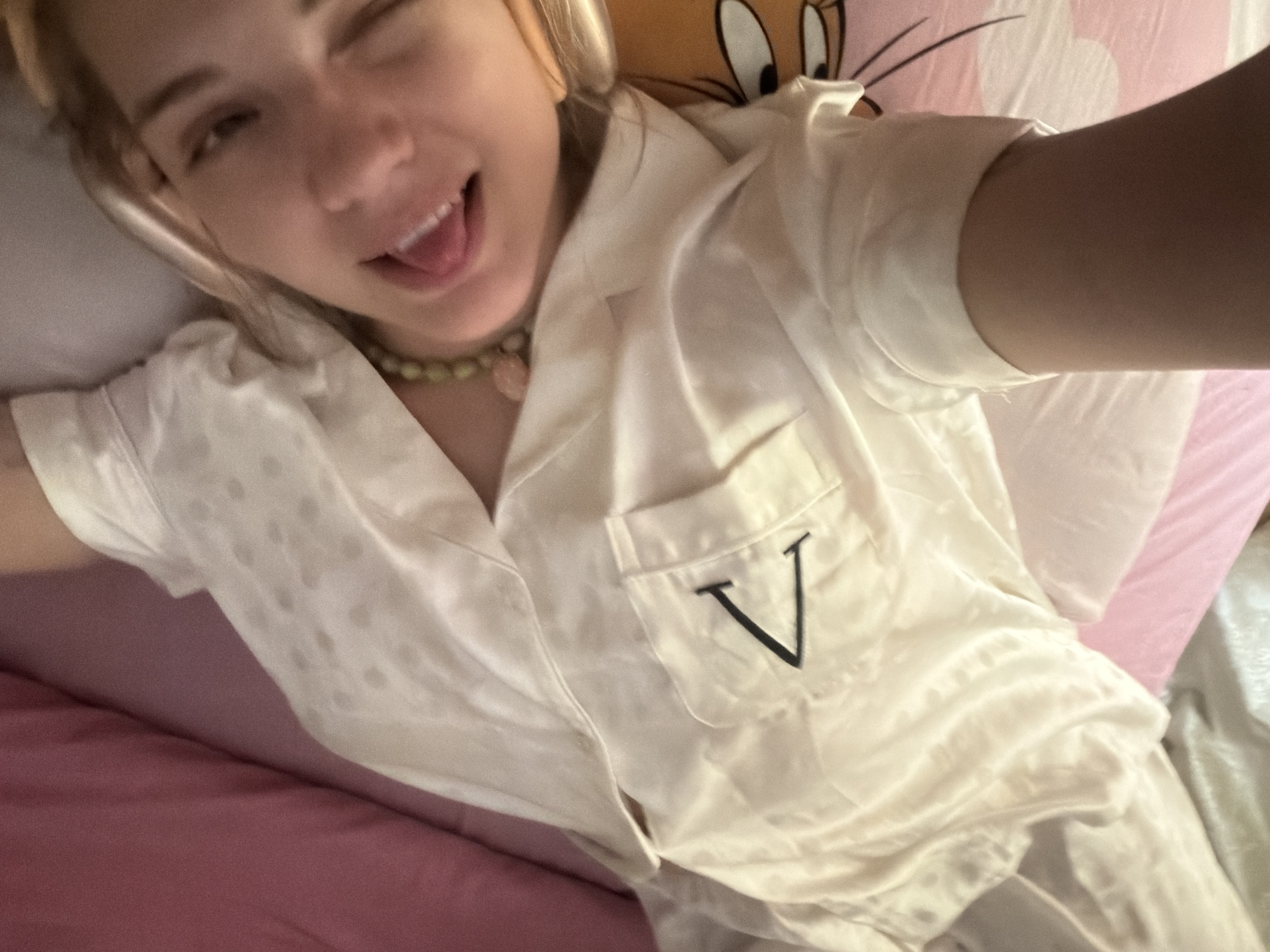 A fair-skinned person with blond hair is lying on a pink bed in what appears to be an indoor location, possibly in Большая Шильна, Russia. The person is wearing a white satin pajama top with a black "V" embroidered on the pocket, and a green beaded necklace. The person is winking and sticking out their tongue. Behind the person is a pillow with a cartoon character on it.