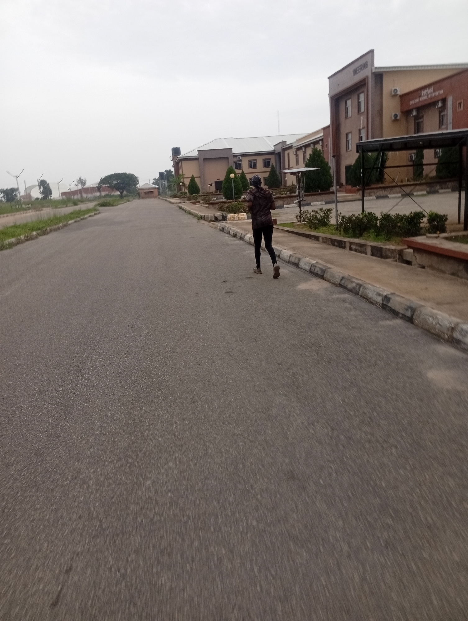 The image shows a person jogging on a paved road in front of a modern building complex. The building, possibly an educational or administrative institution, has multiple stories and features a prominent sign that reads "ENGINEERING". There are trees and landscaping around the buildings. The road appears to be a campus road, with a sidewalk and curb running alongside it. In the background, there are more buildings and some vegetation, along with what appear to be wind turbines in the distance. The sky is overcast, suggesting a cloudy day. The overall impression is of a structured, possibly academic, environment. The location is identified as the city of Jos, Nigeria.