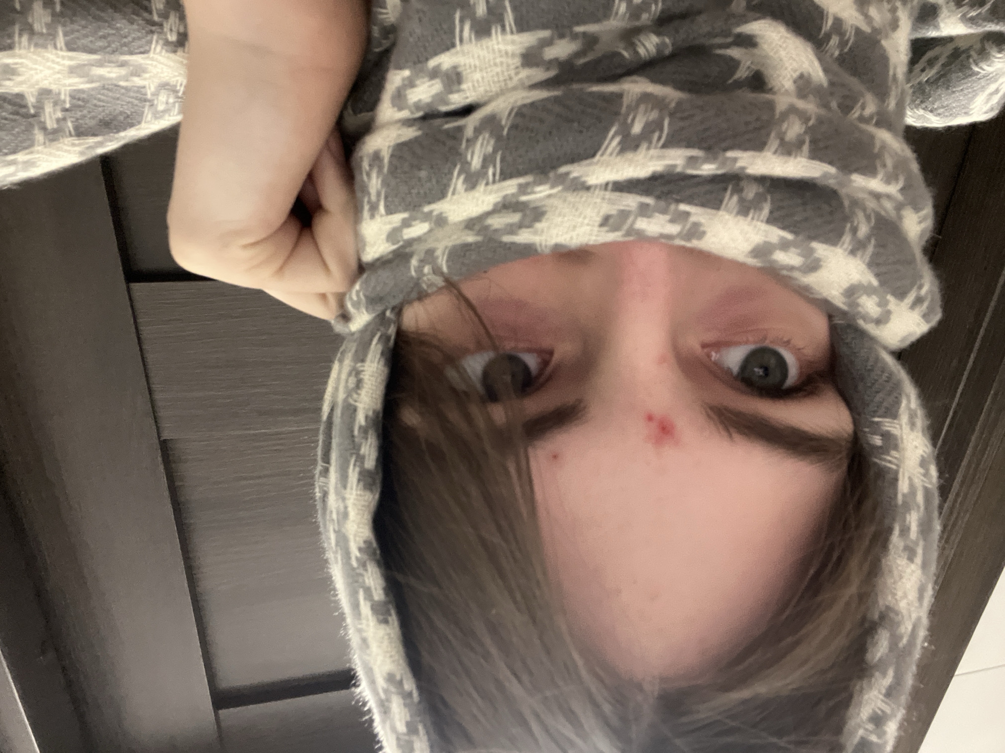 This close-up portrait shows a person's head and upper body, inverted relative to a standard upright orientation. The subject, appearing young, looks directly into the camera with dark grey or hazel eyes. Dark brown hair frames the face, partially obscuring the left side. A grey and off-white patterned scarf or hooded garment, featuring a houndstooth-like design, is wrapped around their head, held in place by a visible hand on the left side. A small red blemish is noticeable on the forehead, just above the nose bridge. The person's expression is neutral and direct. The background is a dark, possibly wooden surface with a visible grain, suggesting an indoor setting. There are no specific location cues visible within the image to indicate Saint Petersburg, Russia, nor any discernible text. The time of day and weather are indeterminate.