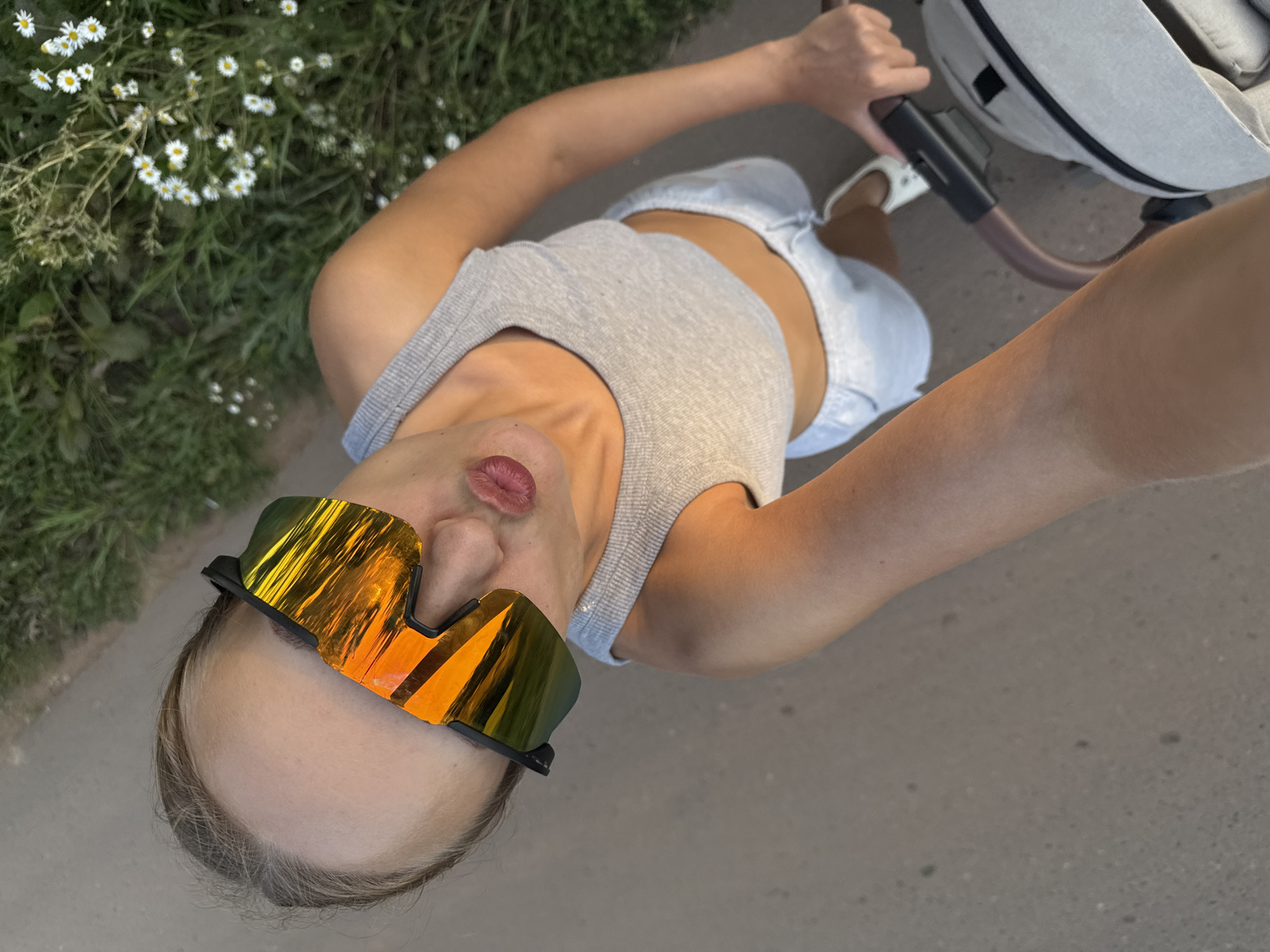 A high-angle selfie captures a person's face, partially obscured by reflective sunglasses with orange and yellow lenses, as they pout their lips. The person is wearing a grey ribbed tank top and light-colored shorts, and their arms are visible. They appear to be holding the handlebars of a stroller. The background shows a paved surface and a grassy area with small white flowers. The overall scene suggests an outdoor activity, possibly a walk or outing, in a natural or park-like setting.