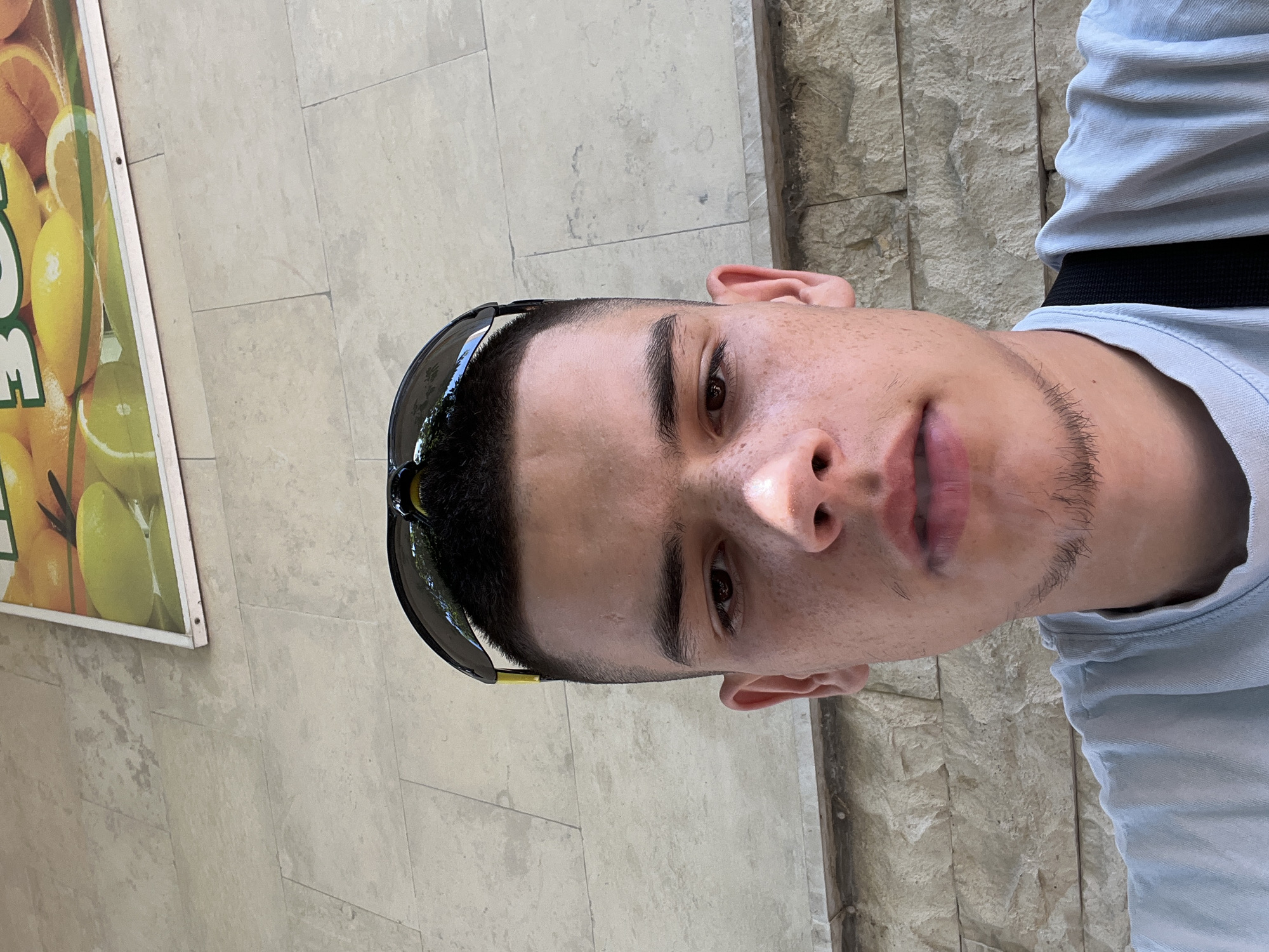 A close-up, selfie-style shot features a young male with short dark hair, sunglasses perched on his head, and freckles across his nose and cheeks. He wears a light blue t-shirt. Behind him, a textured stone wall is visible, with a sign displaying images of citrus fruits, specifically lemons and oranges, mounted above it. The lighting suggests it is daytime. The scene appears to be outdoors. The sign's content suggests a commercial establishment related to fruit or beverages, possibly a cafe or juice bar. There is no direct indication of the city of Karlovo, Bulgaria, within the image itself.
