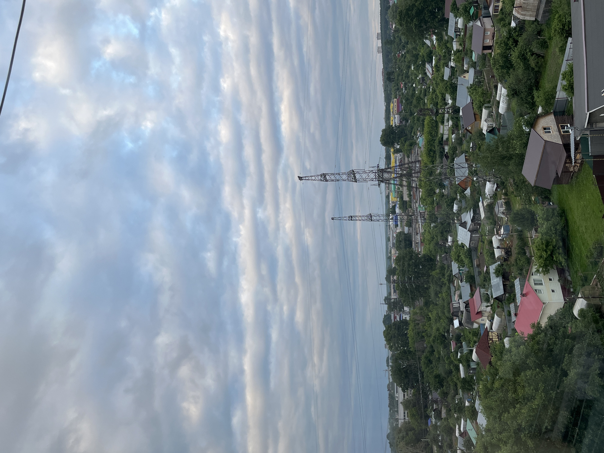 The image shows an aerial view of a semi-urban or suburban area with many small houses and gardens. There are two large electrical transmission towers in the middle ground, one taller than the other. Power lines stretch across the scene. The sky is overcast with layered clouds. The landscape is lush with many trees and green vegetation. Some of the houses have red roofs, while others have gray or brown roofs. There are also several greenhouses visible. In the distance, there are more buildings, some appearing to be industrial or taller residential structures, and a few more transmission towers. The overall impression is of a densely populated residential area surrounded by natural greenery, with significant infrastructure from power transmission.