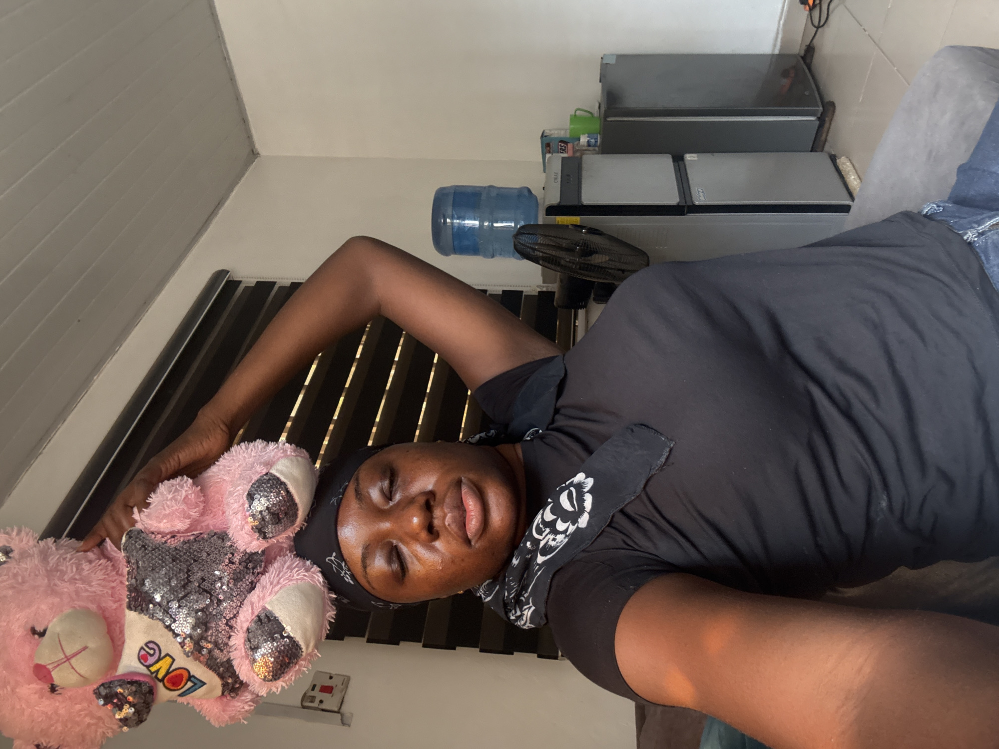 A dark-skinned woman with a black headscarf and a patterned black scarf around her neck is lying down, holding a pink teddy bear. The teddy bear has "LOVE" written on its foot. The woman is lying against a window with blinds. A water cooler and two portable refrigerators are visible in the background. Based on the presence of the water cooler, refrigerators, and the general appearance of the room, the location appears to be a domestic interior. Without further information, it is not possible to determine if the location is in the city of Abuja, Nigeria.