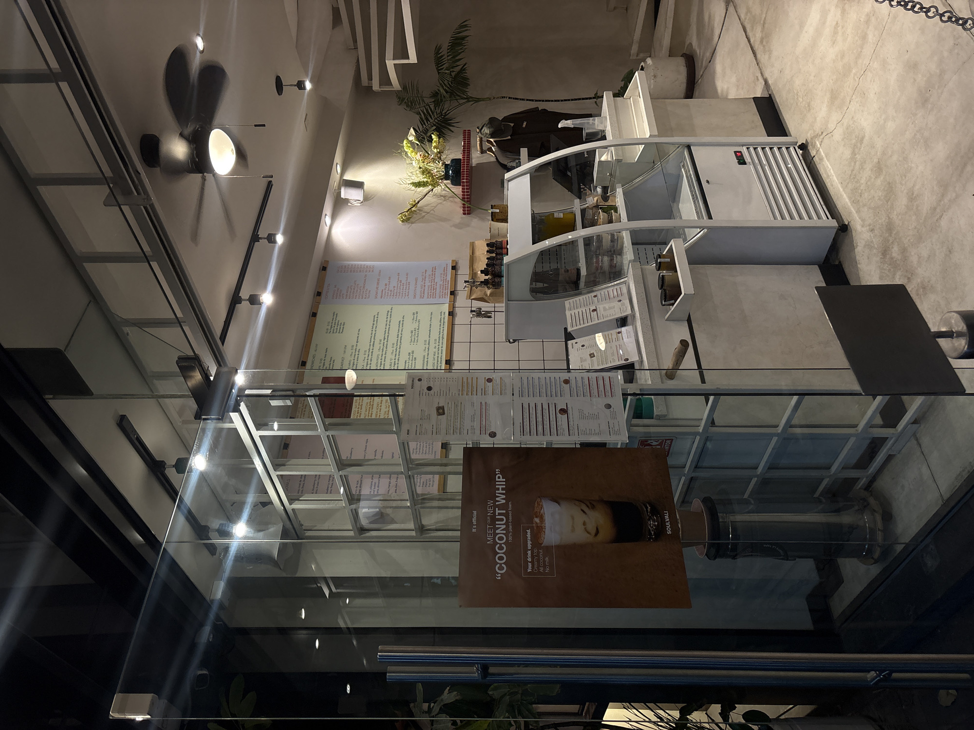 Here's a detailed description of the image:
**Scene:** The photograph shows the interior of a cafe or restaurant, possibly in Puebla, Mexico. The dominant features are the service counter, a display case, and the surrounding decor.
**Objects:**
* **Service Counter:** A prominent white service counter with glass display shelves is visible. The shelves hold food items.
* **Menu Boards:** Behind the counter, a series of menus are hung on the wall, listing various food and drink options.
* **"Coconut Whip" Advertisement:** A poster advertising "Coconut Whip" (a plant-based foam topping) is displayed on a glass barrier, possibly at the entrance or front of the counter. The poster has details about the product and the brand name "SOULVALI".
* **Ceiling Fan and Lighting:** A ceiling fan is mounted on the ceiling. Track lighting fixtures are also present.
* **Decor:** Decorative elements include plants, possibly a palm tree, and other floral arrangements.
**Environment:**
* The interior design appears modern and clean.
* The floor is a light, possibly concrete, color.
* The lighting is a combination of natural and artificial.
**Location:**
* The brand SOULVALI is known to be in the city of Puebla, Mexico.