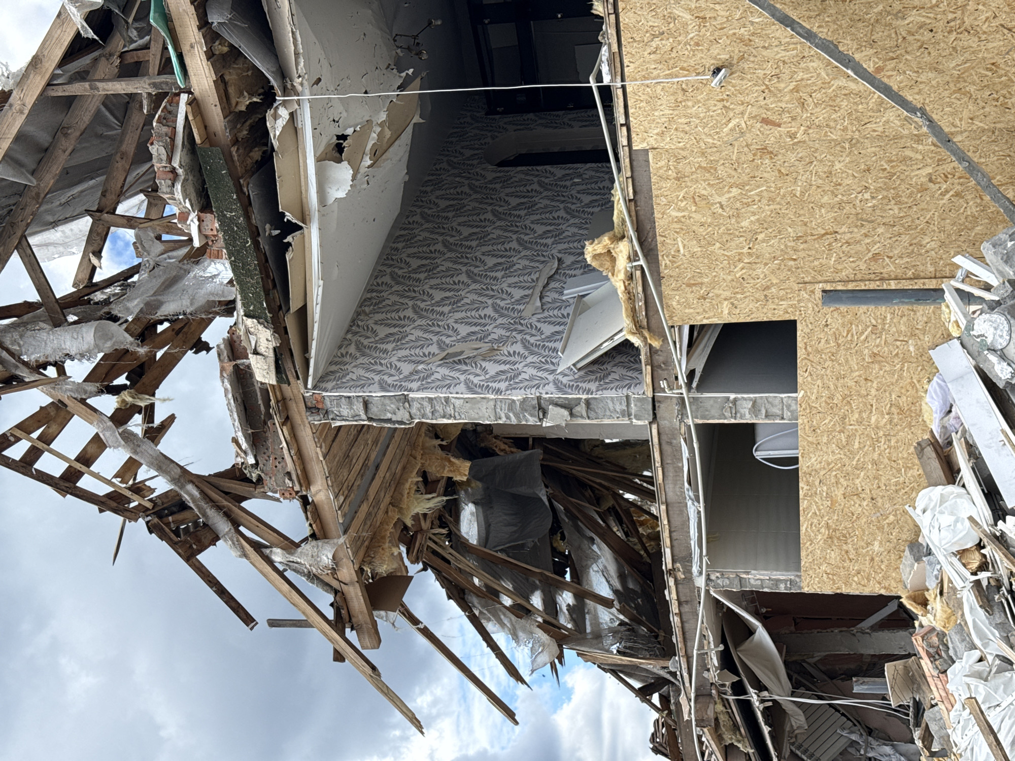The image depicts the aftermath of an explosion or significant structural damage to a multi-story building in Kyiv, Ukraine. The roof and upper floors have been severely compromised, with exposed wooden beams, debris, insulation, and shattered building materials scattered throughout. The outer walls show signs of blast damage, with some sections appearing intact while others are missing or partially collapsed. The interior of the building is visible in some areas, revealing wallpaper, exposed wiring, and remnants of household items. The lower levels appear to be partially obscured by rubble and construction materials, suggesting ongoing cleanup or stabilization efforts. The sky above is overcast, with scattered clouds. There are no people visible in the image. The overall scene conveys destruction and damage.
