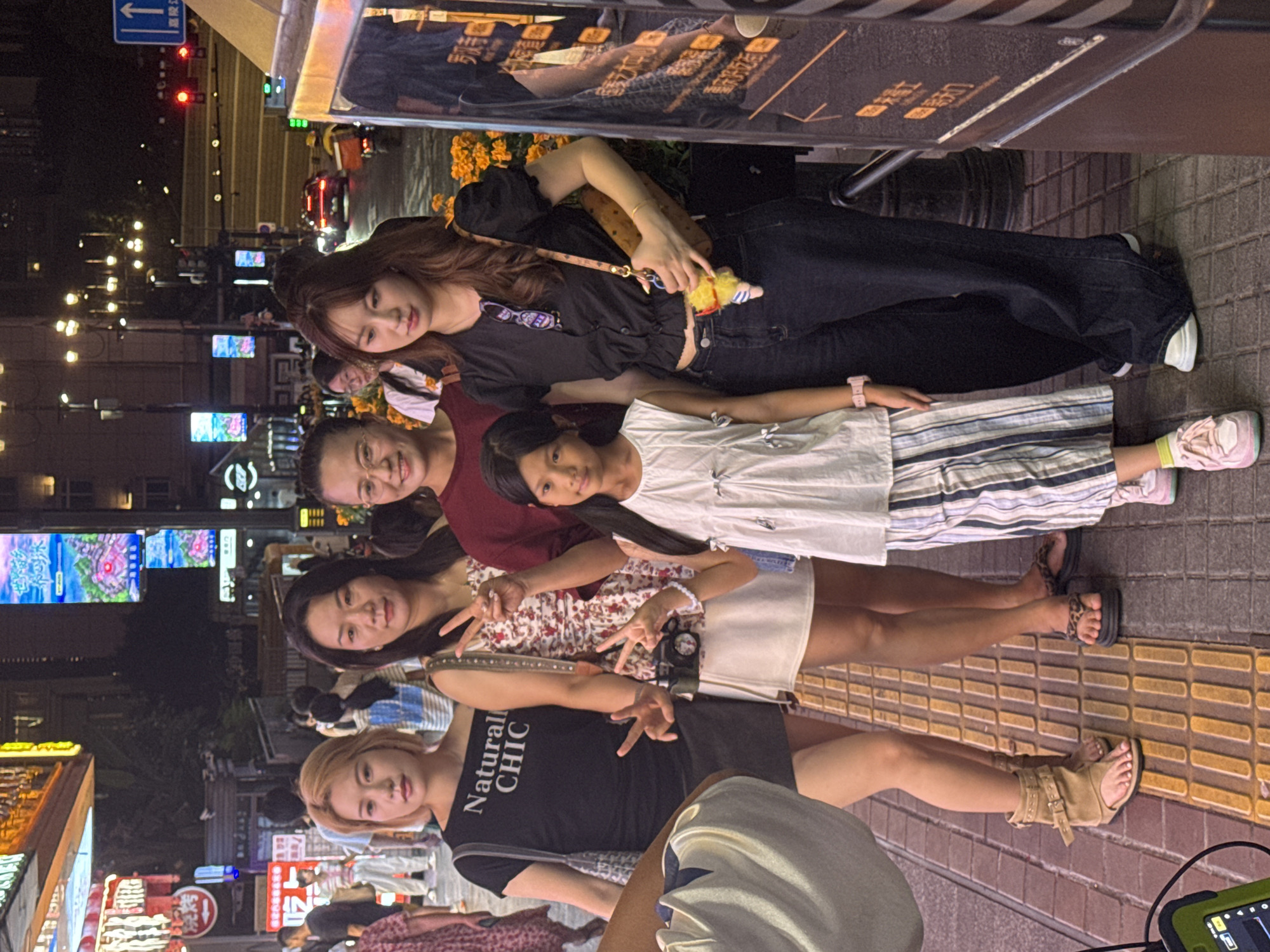 In Yuzhong District, China, at night, five people are posing for a photo on a sidewalk. From left to right, a woman with short blonde hair wears a black off-the-shoulder shirt that reads "Natural CHIC" and a dark skirt. Next to her, a woman with long dark hair wears a floral print dress and a small camera hangs from her shoulder. A young girl with black hair stands in front of a woman wearing a maroon top and glasses. The fifth person, a young woman with long brown hair, wears a black short-sleeved button-up shirt and dark jeans, holding a brown purse. The background features illuminated buildings, streetlights, and passing vehicles, indicating a busy urban environment. The people are casually dressed, and their poses suggest they are friends or family.