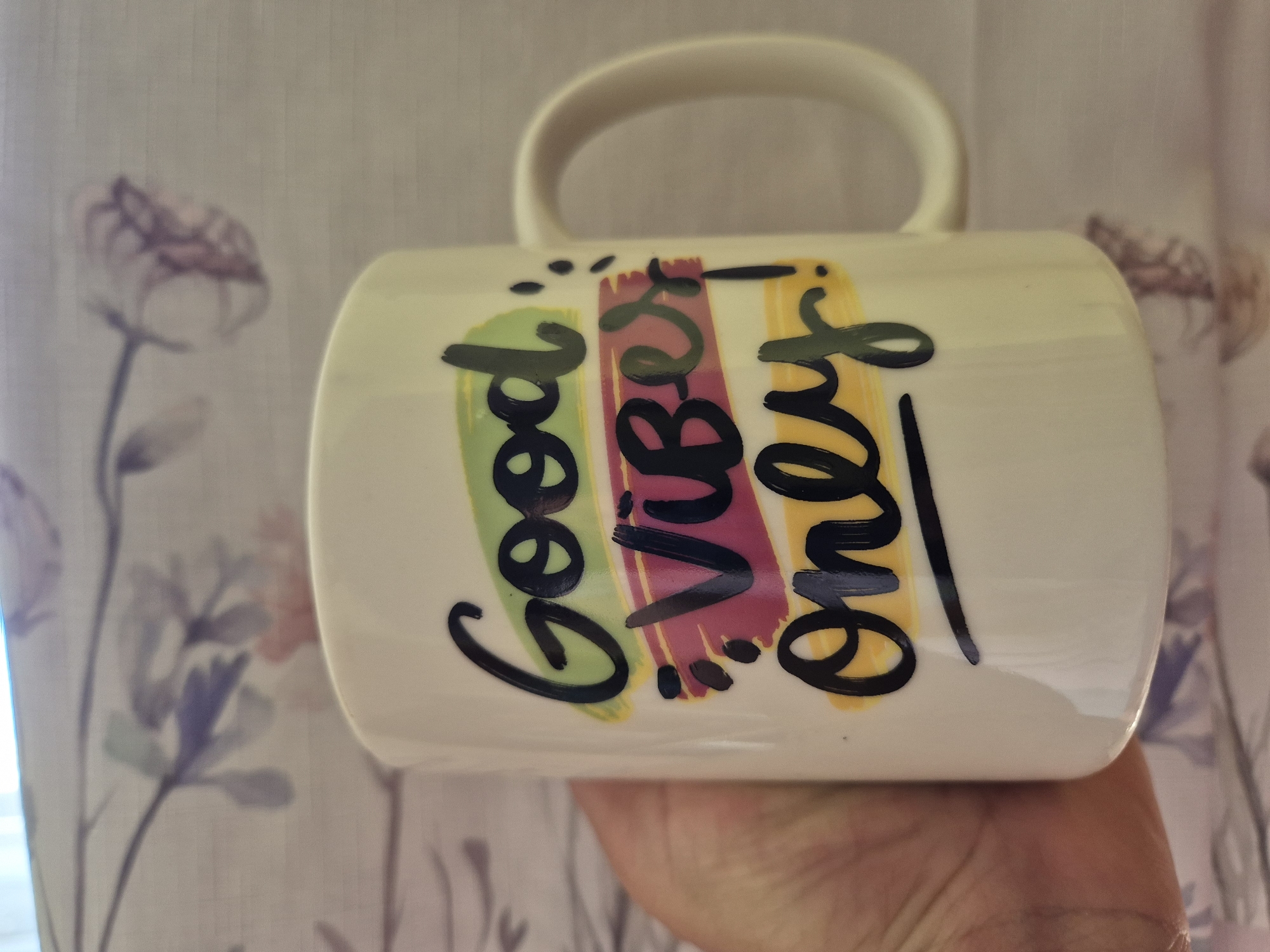 A white mug is held in someone's hand. The mug has a handle and is decorated with the words "Good Vibes Only" in black cursive writing with colorful backgrounds of green, pink, and yellow. The background is a floral print curtain. The location is unknown.
