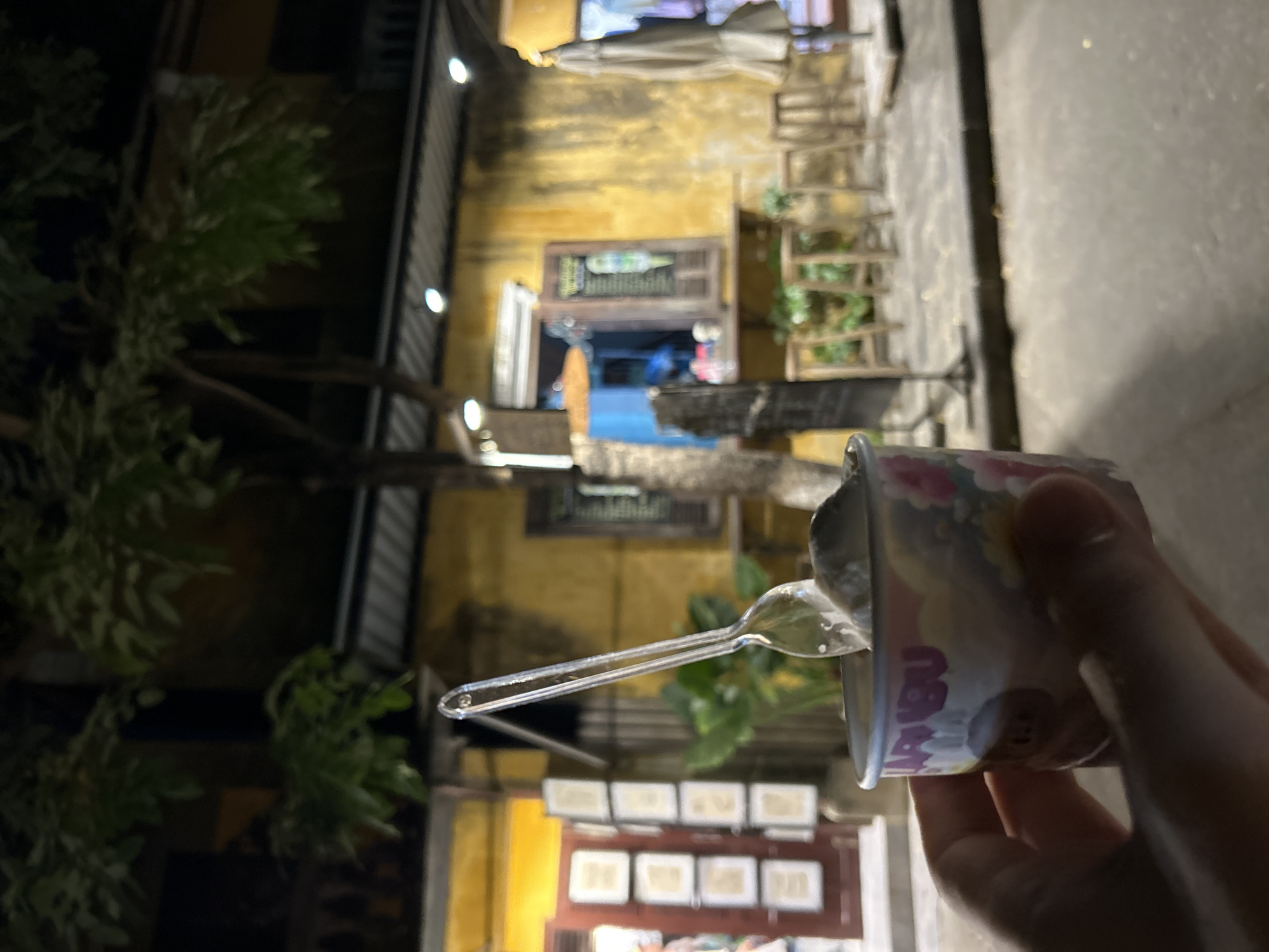 A first-person perspective shot shows a hand holding a small cup of ice cream with a plastic spoon in it. The cup is colorful with cartoonish designs of flowers and animals, and the word "MARIBU" is visible on it. The background features a dimly lit street scene in Hoi An, Vietnam, at night. A weathered, yellow building with a small open window is behind the ice cream cup. The window displays a menu board with an image of ice cream. Several wooden stools are lined up in front of the building, and a closed umbrella stands to the right. The scene is set outdoors on a paved surface, with trees and foliage overhead. The lighting suggests it is evening or night.